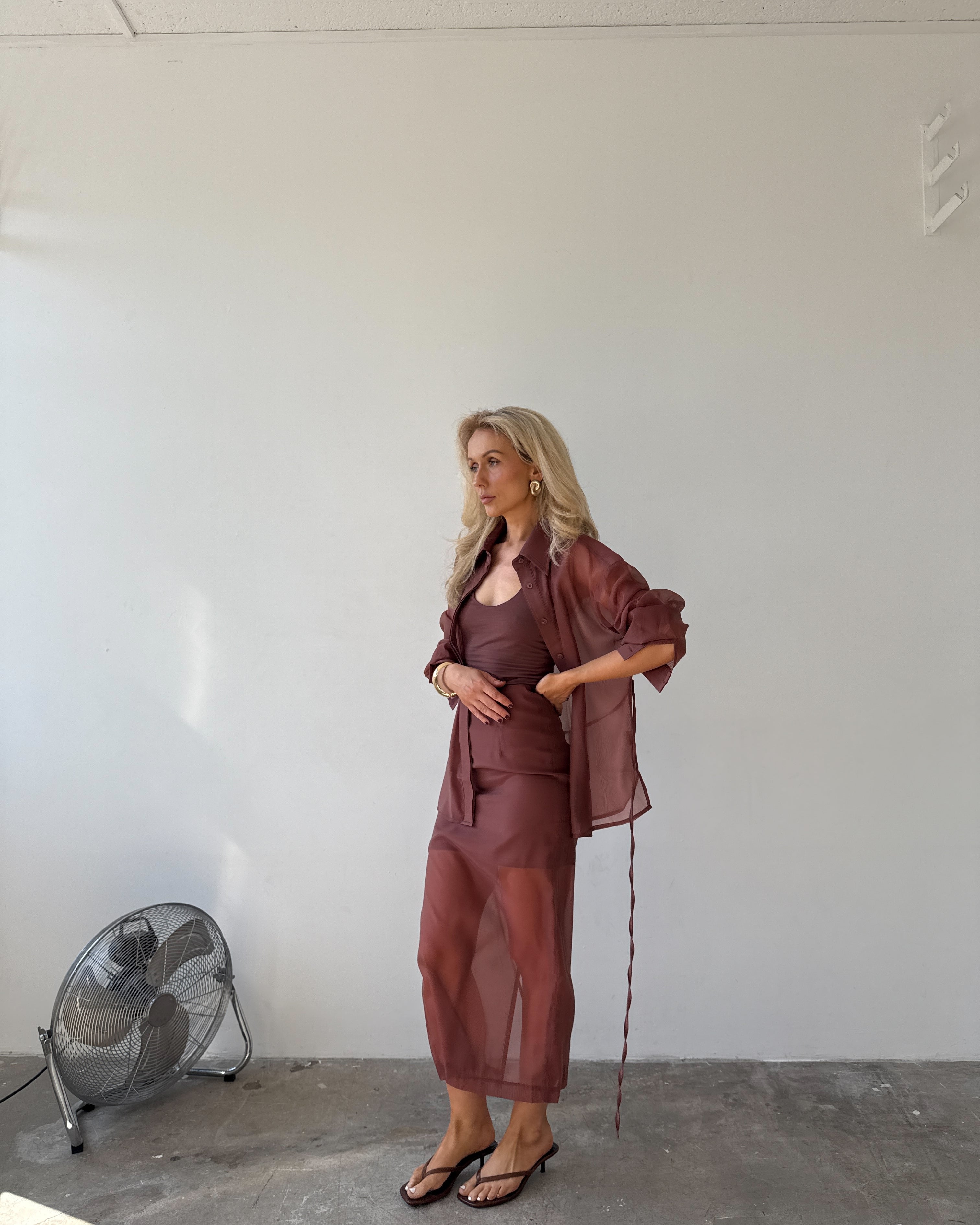 Model in a red-brown outfit standing in a minimalistic room with a fan