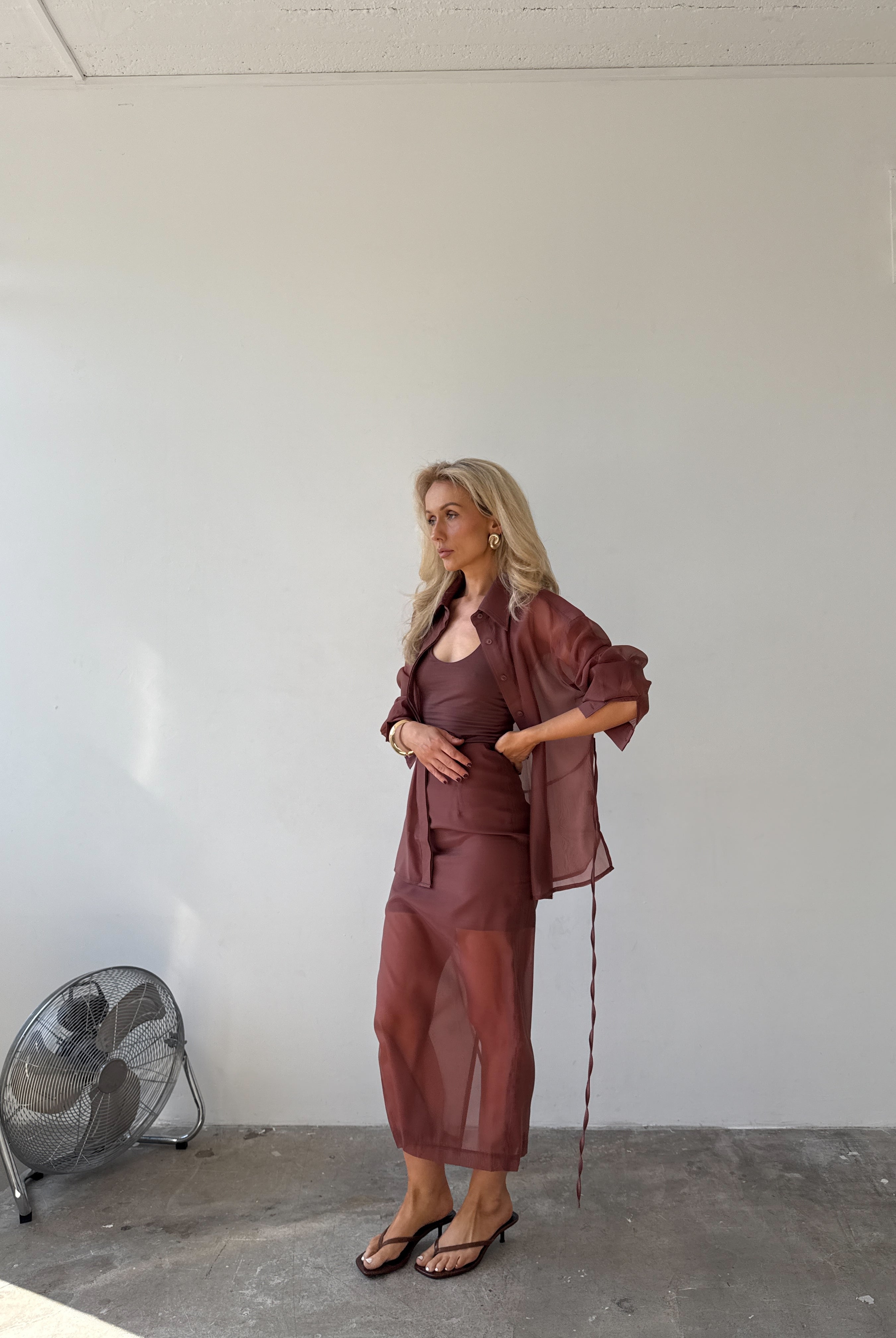 Model in a red-brown outfit standing in a minimalistic room with a fan