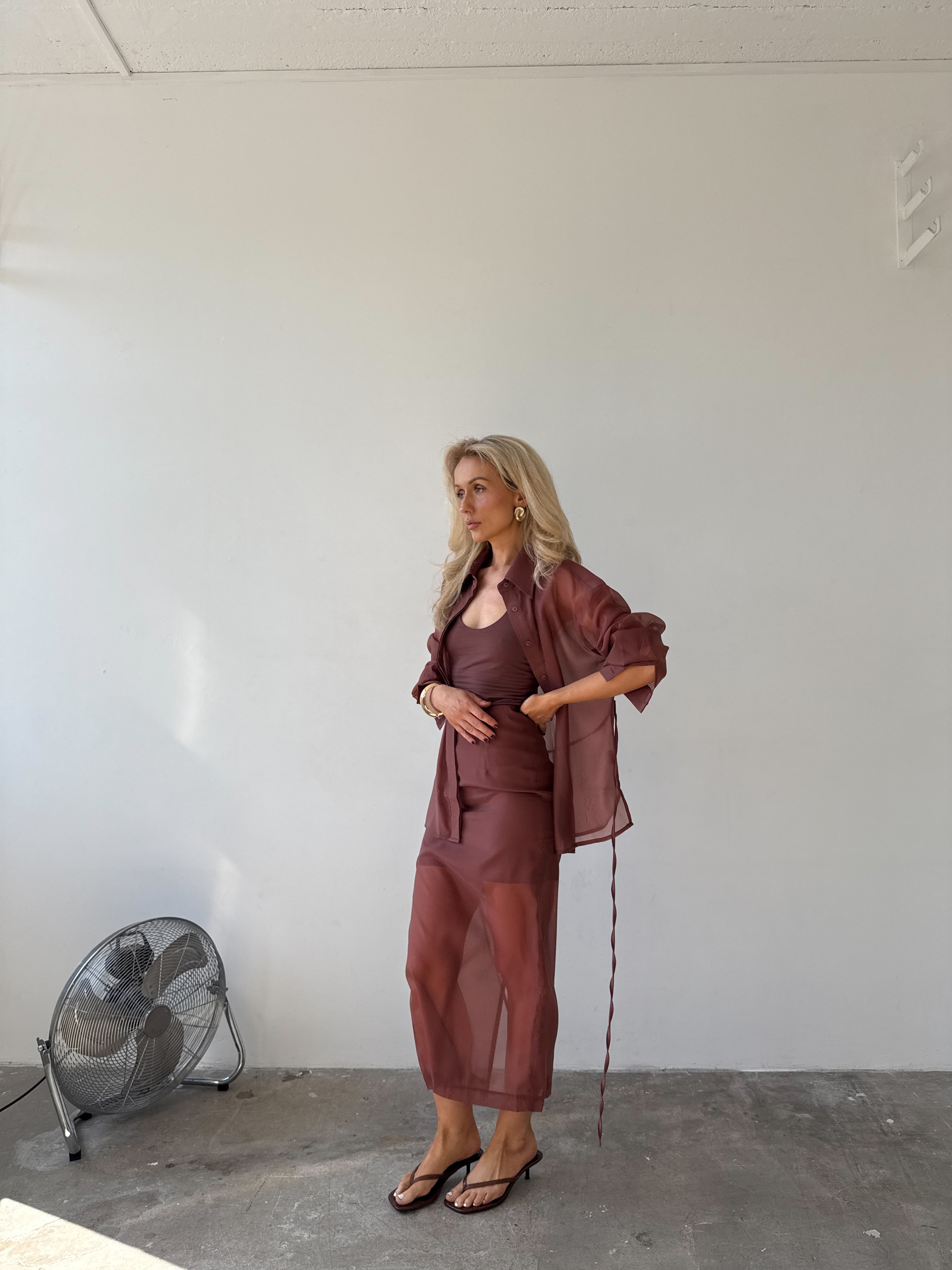 Model in a red-brown outfit standing in a minimalistic room with a fan