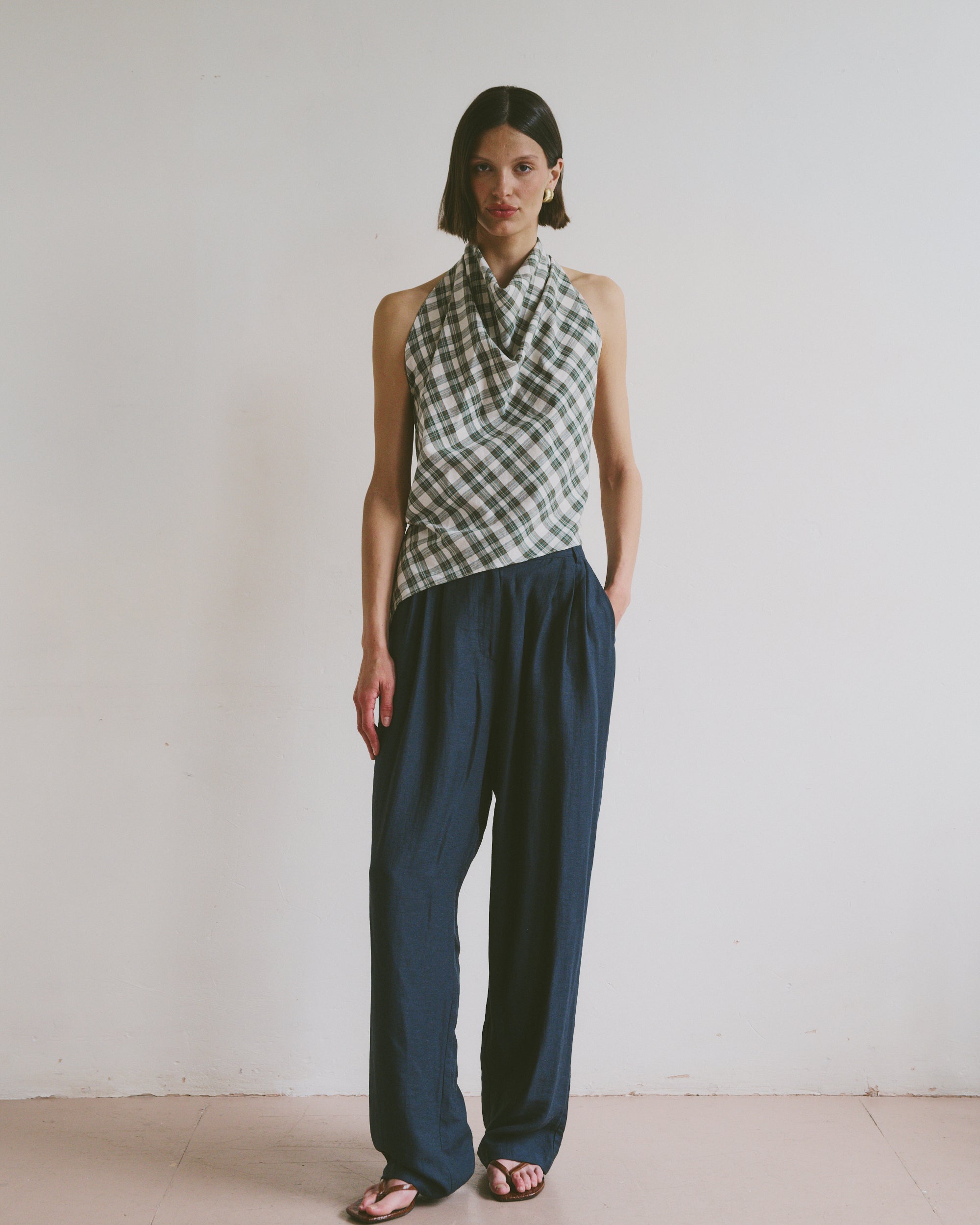 Model wears ink blue, flowy linen trousers styled with a checkered, high-neck sleeveless top and black sandals