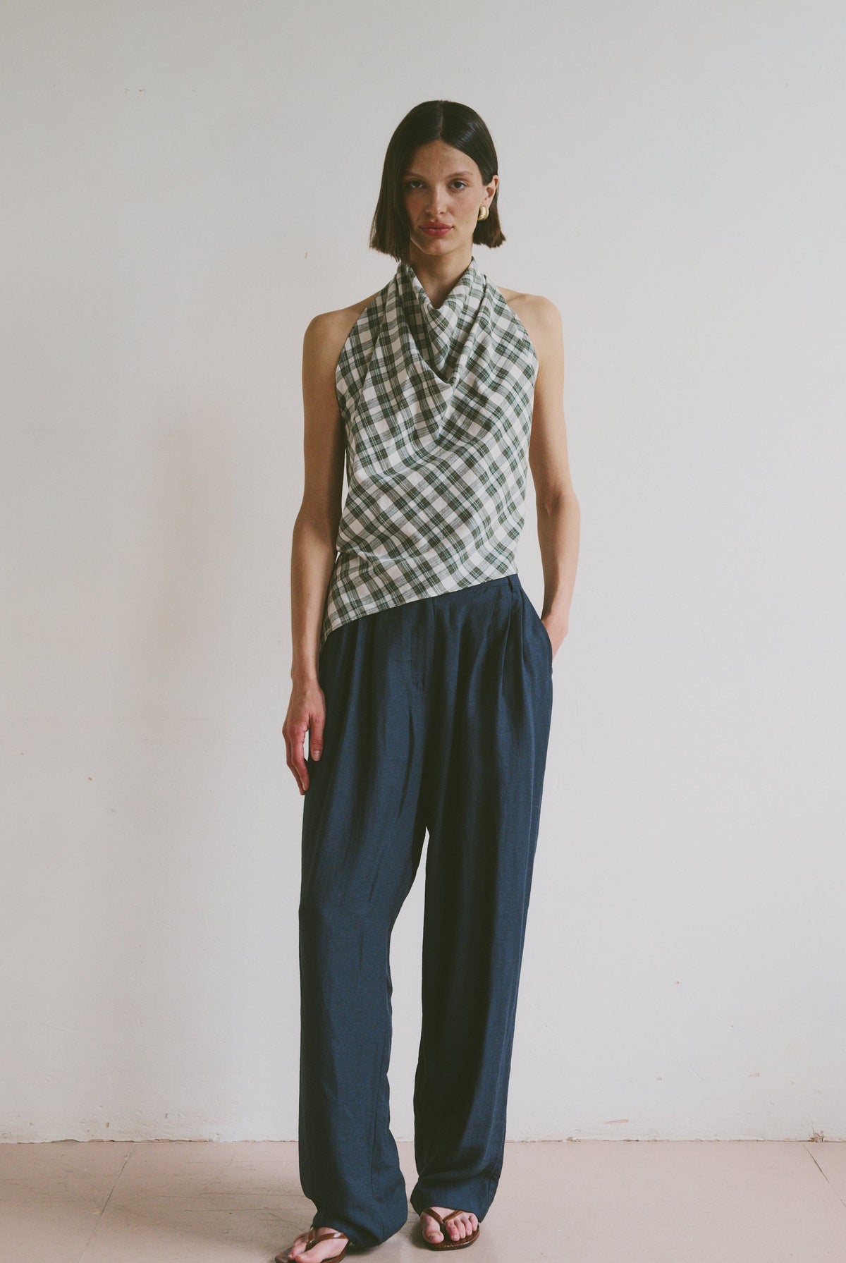 Model wears ink blue, flowy linen trousers styled with a checkered, high-neck sleeveless top and black sandals
