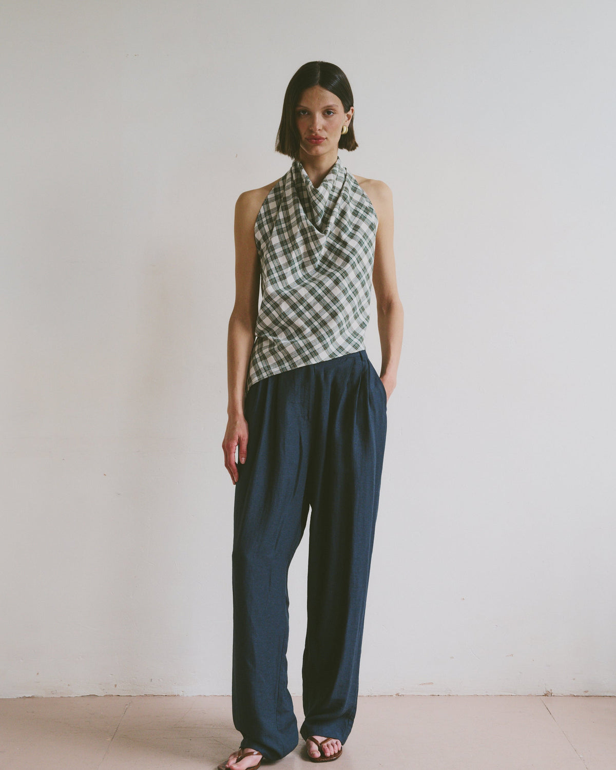 Model wears ink blue, flowy linen trousers styled with a checkered, high-neck sleeveless top and black sandals