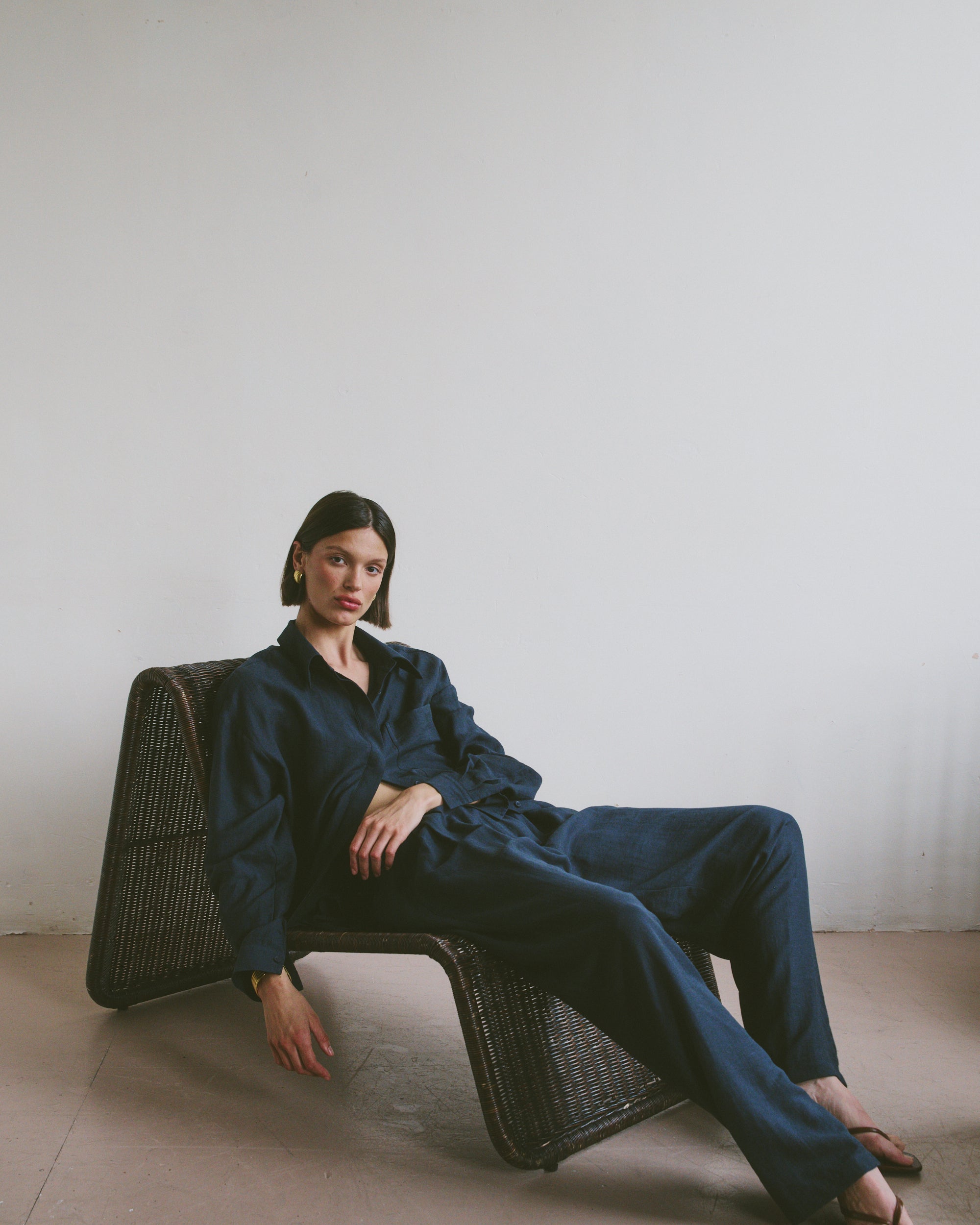 Model in a navy blue outfit sitting on a wicker chair in a relaxed position against a plain wall