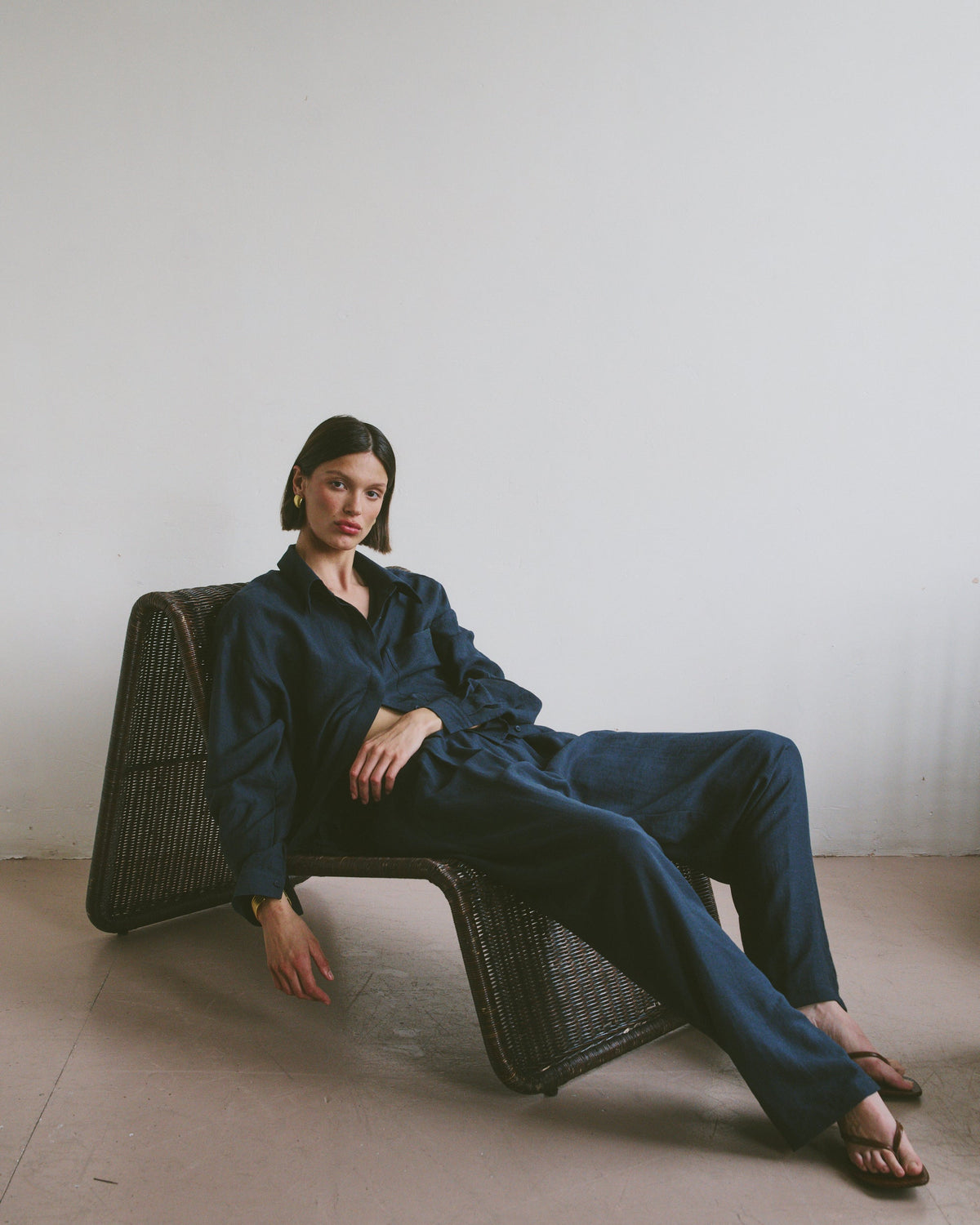 Model in a navy blue outfit sitting on a wicker chair in a relaxed position against a plain wall