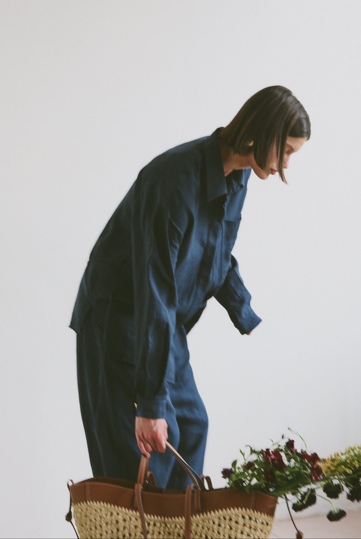 Model wearing a blue outfit holding a woven handbag with flowers and greenery coming out the top