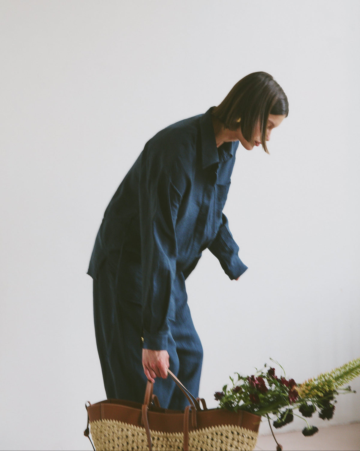 Model wearing a blue outfit holding a woven handbag with flowers and greenery coming out the top