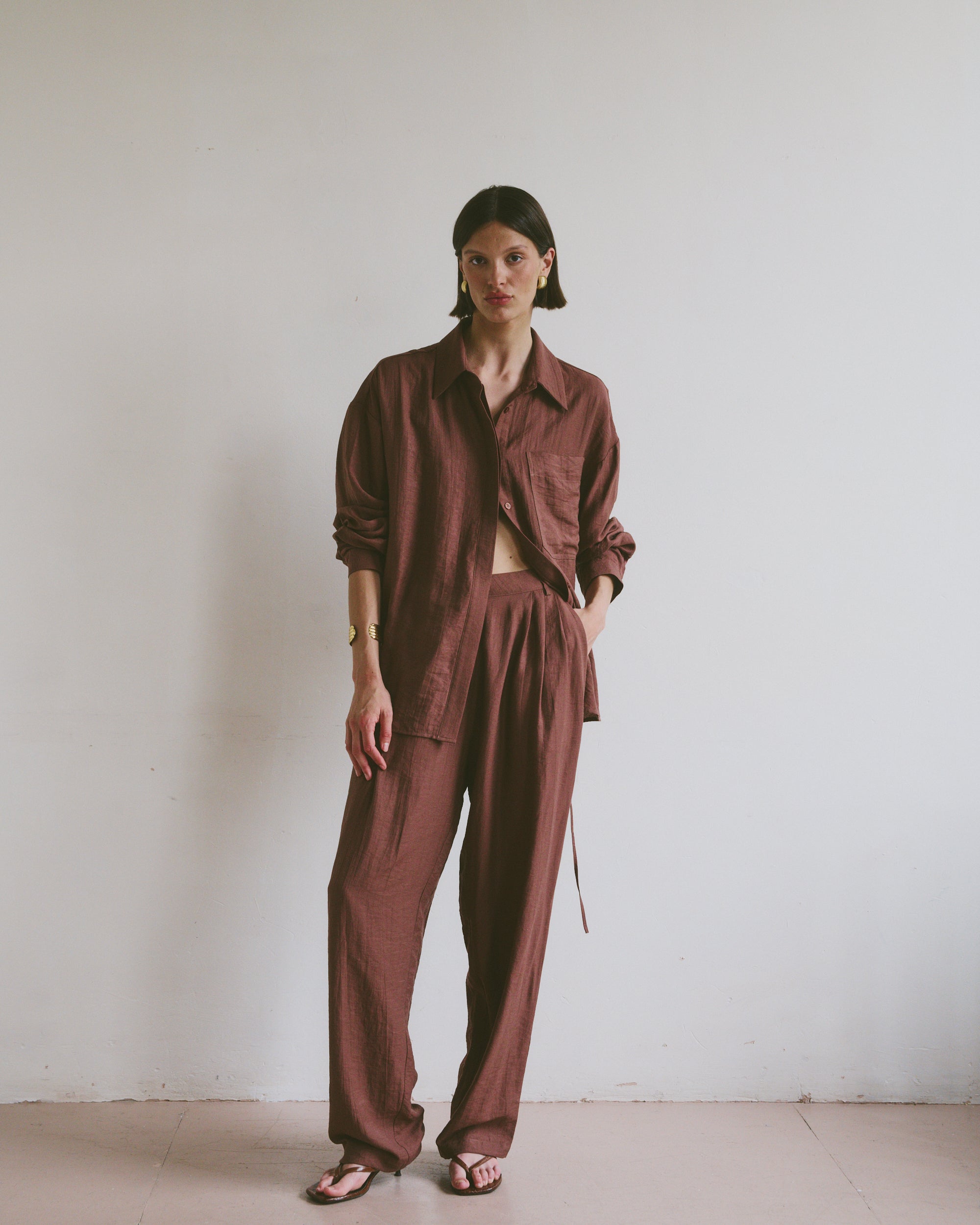 Model wearing a relaxed maroon linen shirt and matching trousers styled with open toe sandals in front of a minimalistic background