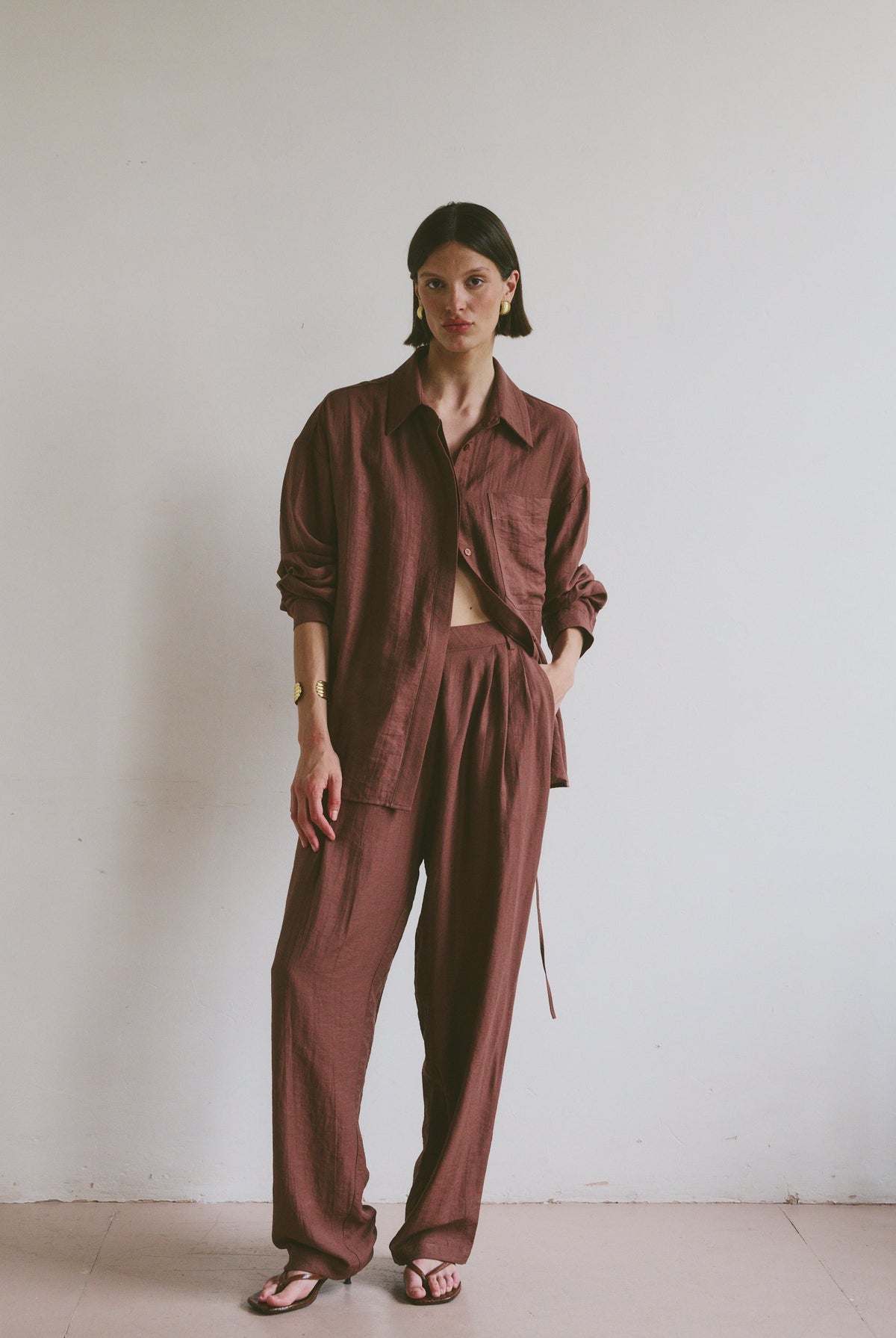 Model wearing a relaxed maroon linen shirt and matching trousers styled with open toe sandals in front of a minimalistic background