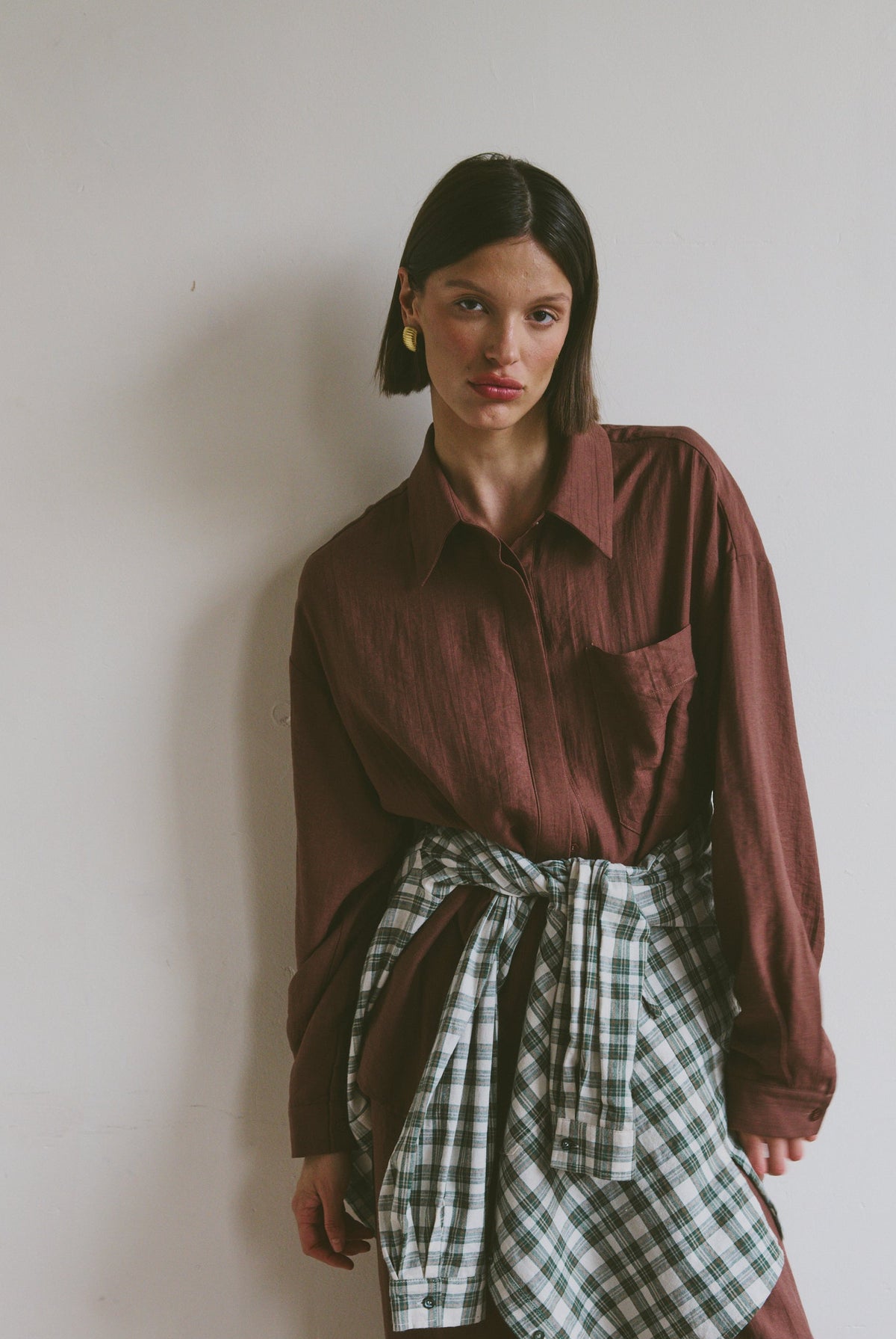 Model wearing a burgundy shirt with matching trousers with a plaid shirt tied around their waist against a plain background