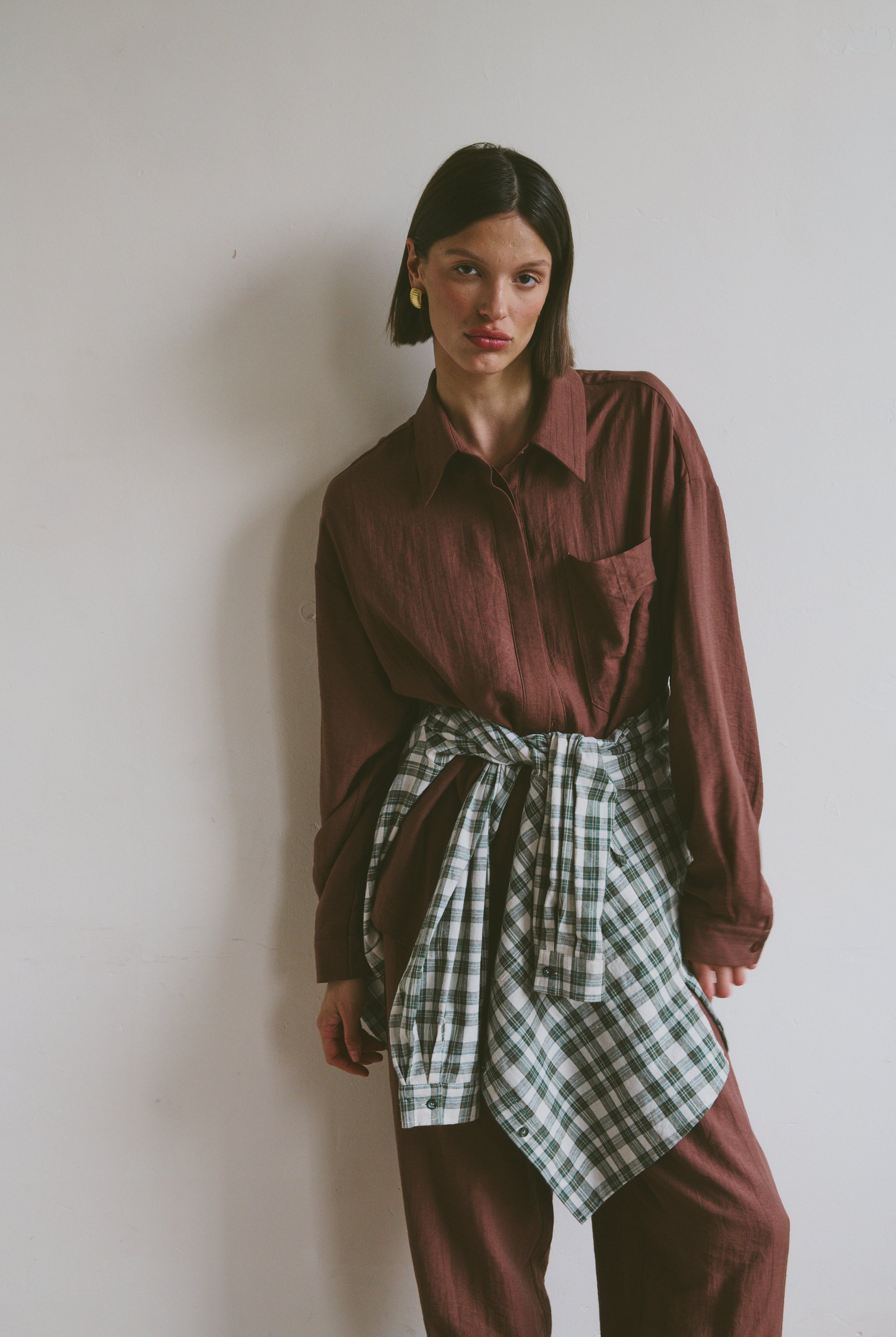 Model wearing a burgundy shirt with matching trousers with a plaid shirt tied around their waist against a plain background