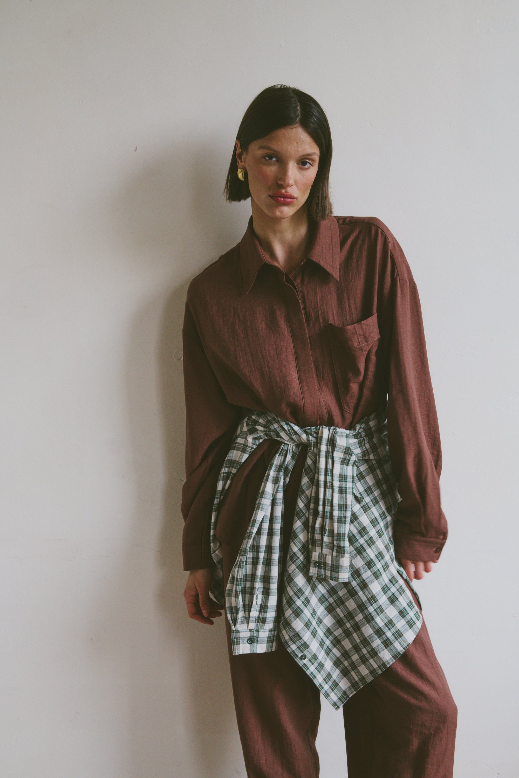 Model wearing a burgundy shirt with matching trousers with a plaid shirt tied around their waist against a plain background