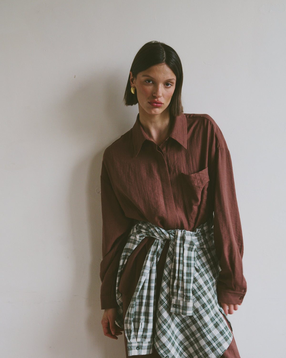 Model wearing a burgundy shirt with matching trousers with a plaid shirt tied around their waist against a plain background