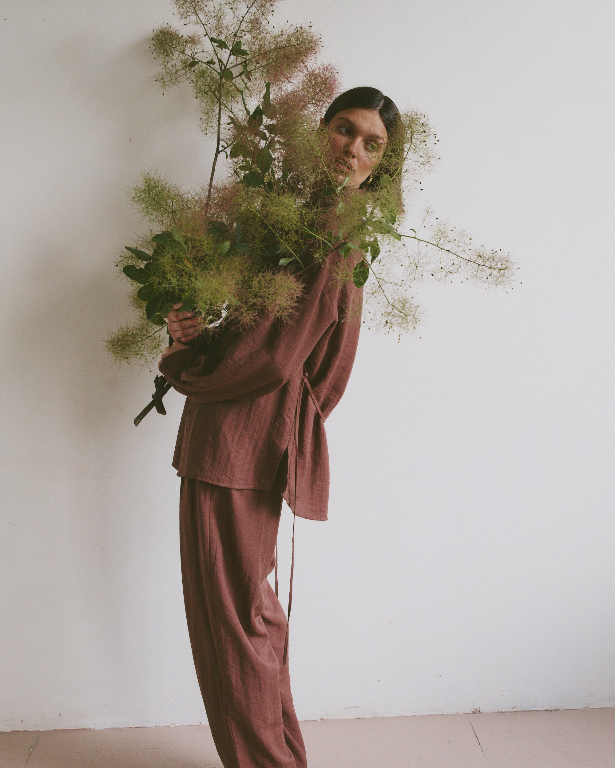 Model in a loose-fitting, mauve-toned outfit holding a large, airy bouquet of greenery, standing against a plain white wall