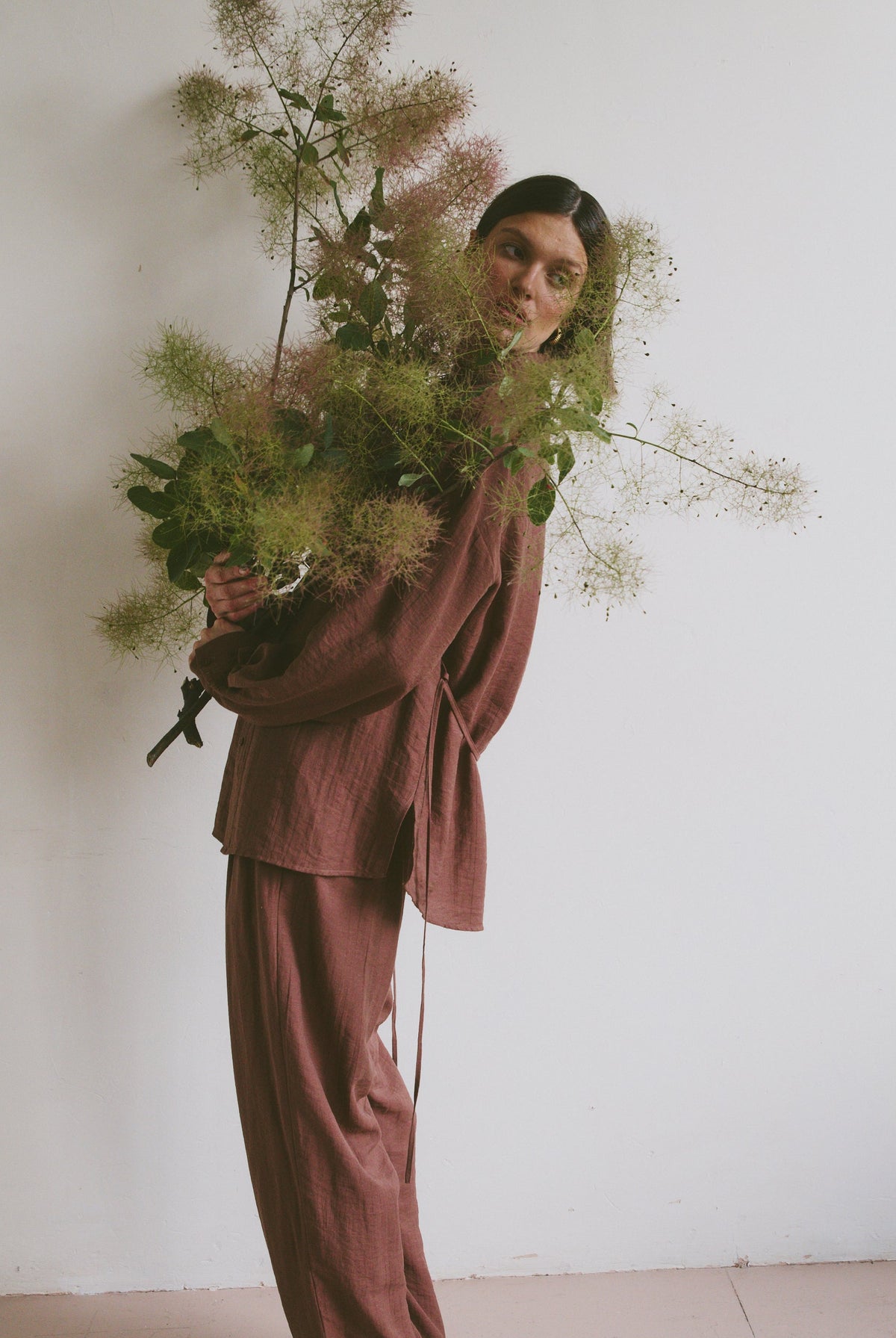 Model in a loose-fitting, mauve-toned outfit holding a large, airy bouquet of greenery, standing against a plain white wall