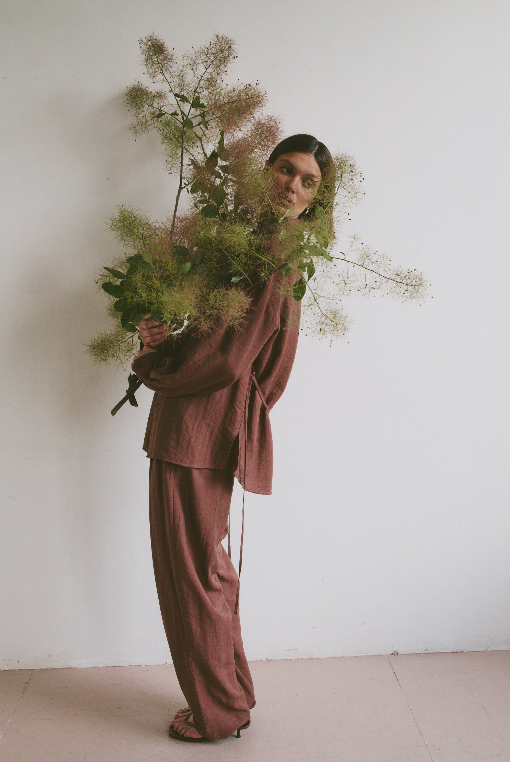 Model in a loose-fitting, mauve-toned outfit holding a large, airy bouquet of greenery, standing against a plain white wall
