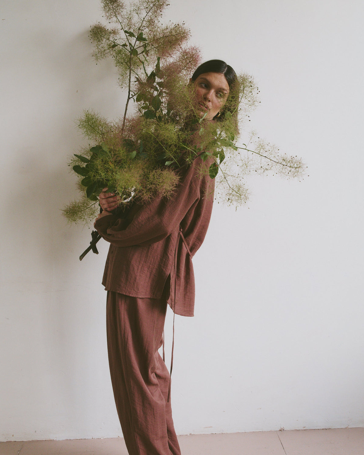 Model in a loose-fitting, mauve-toned outfit holding a large, airy bouquet of greenery, standing against a plain white wall