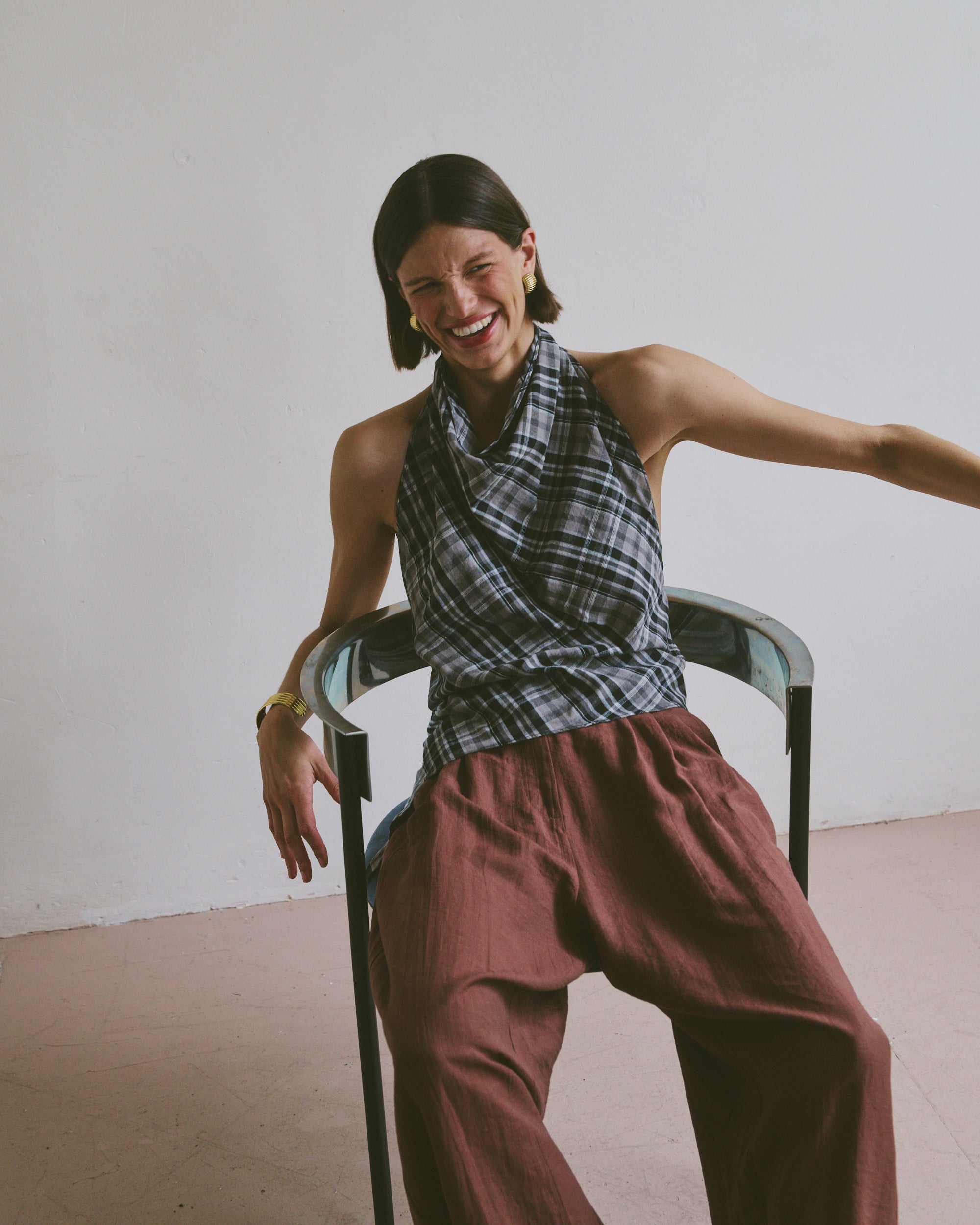 Woman laughing while sitting on a chair wearing a plaid top and burgundy pants against a plain background