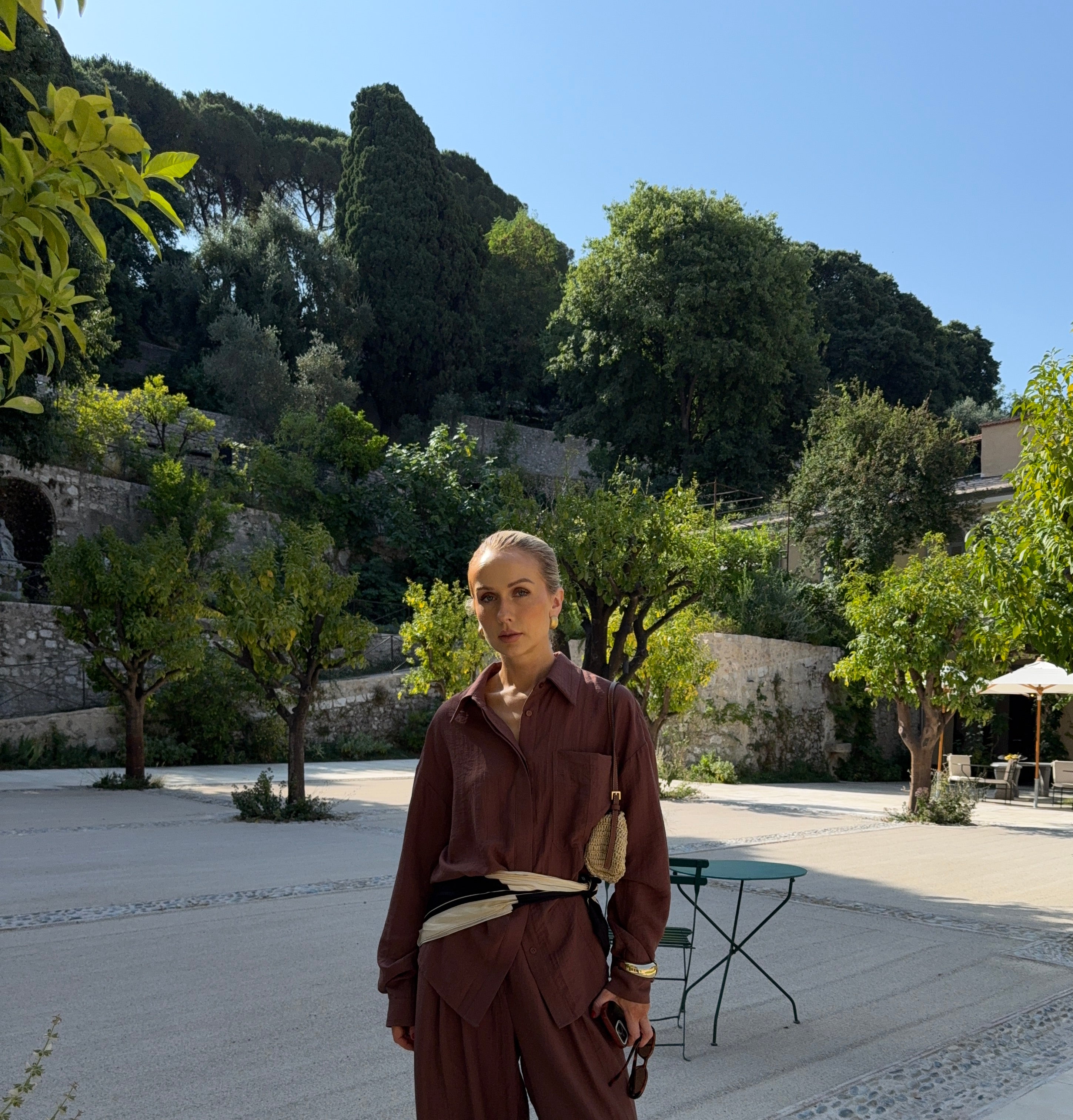 Woman in auburn-brown outfit standing on a paved area with trees and buildings in the background