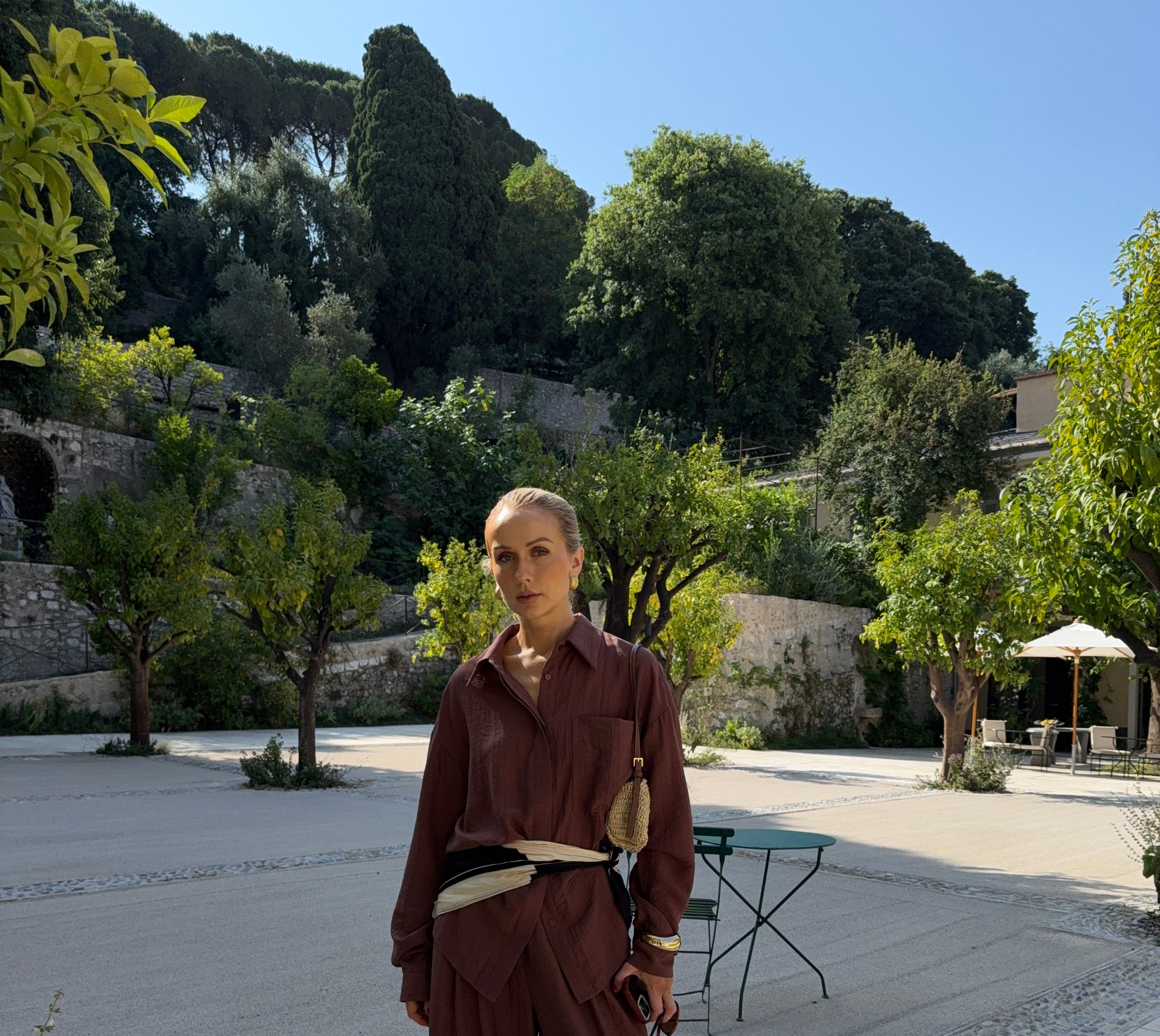 Woman in auburn-brown outfit standing on a paved area with trees and buildings in the background