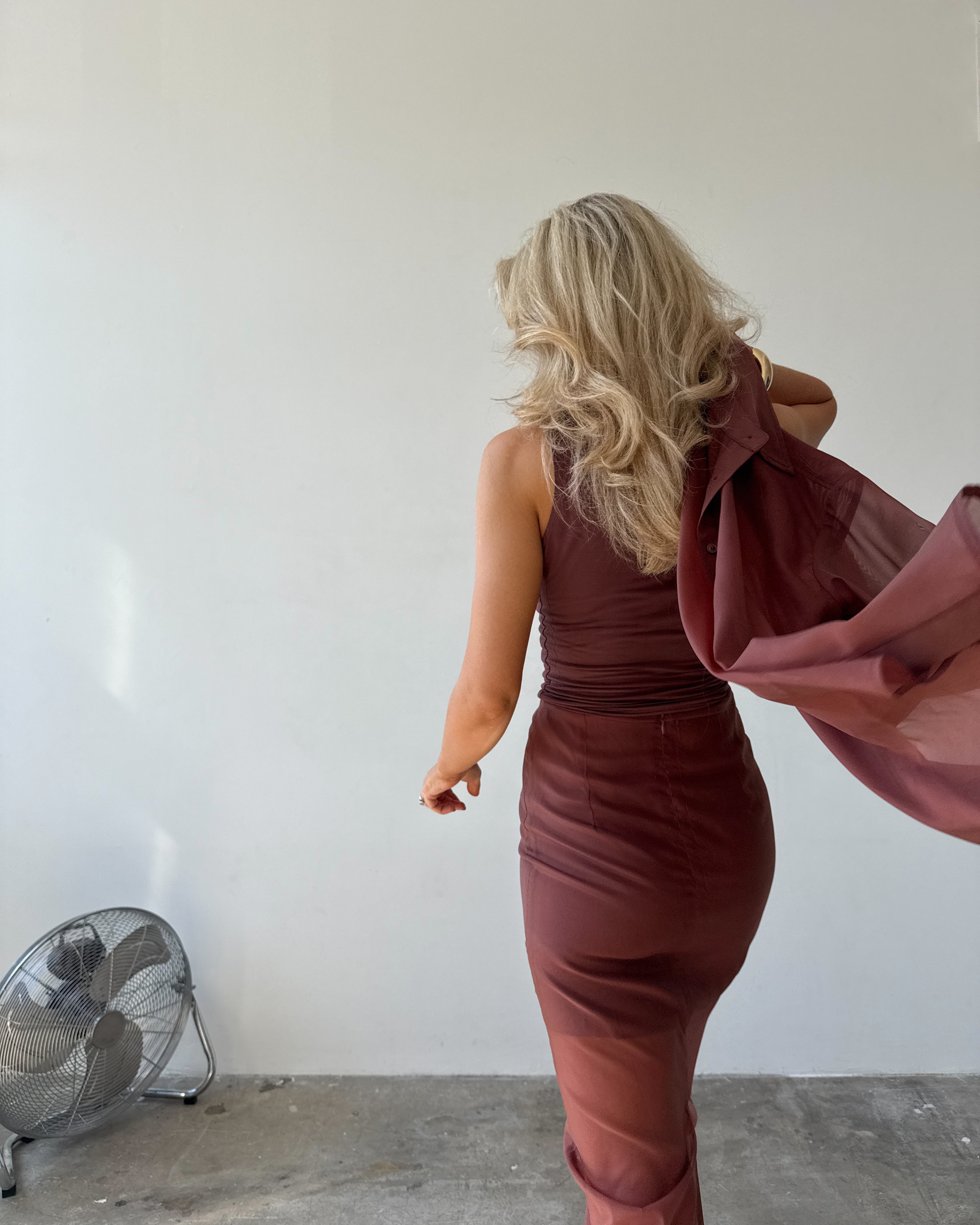 Woman walking away from the camera in a maroon top and skirt holding a matching jacket against a plain wall