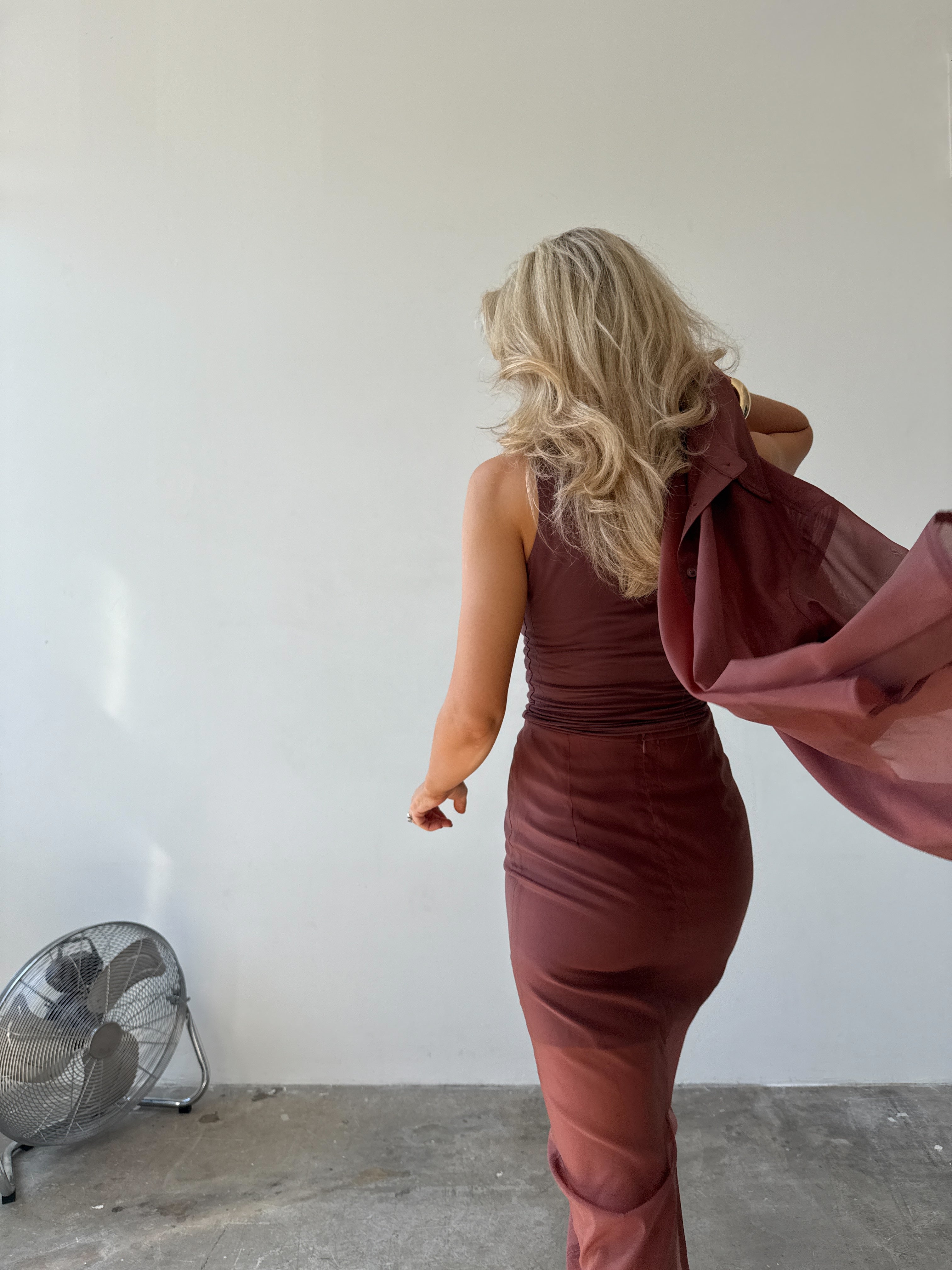Woman walking away from the camera in a maroon top and skirt holding a matching jacket against a plain wall