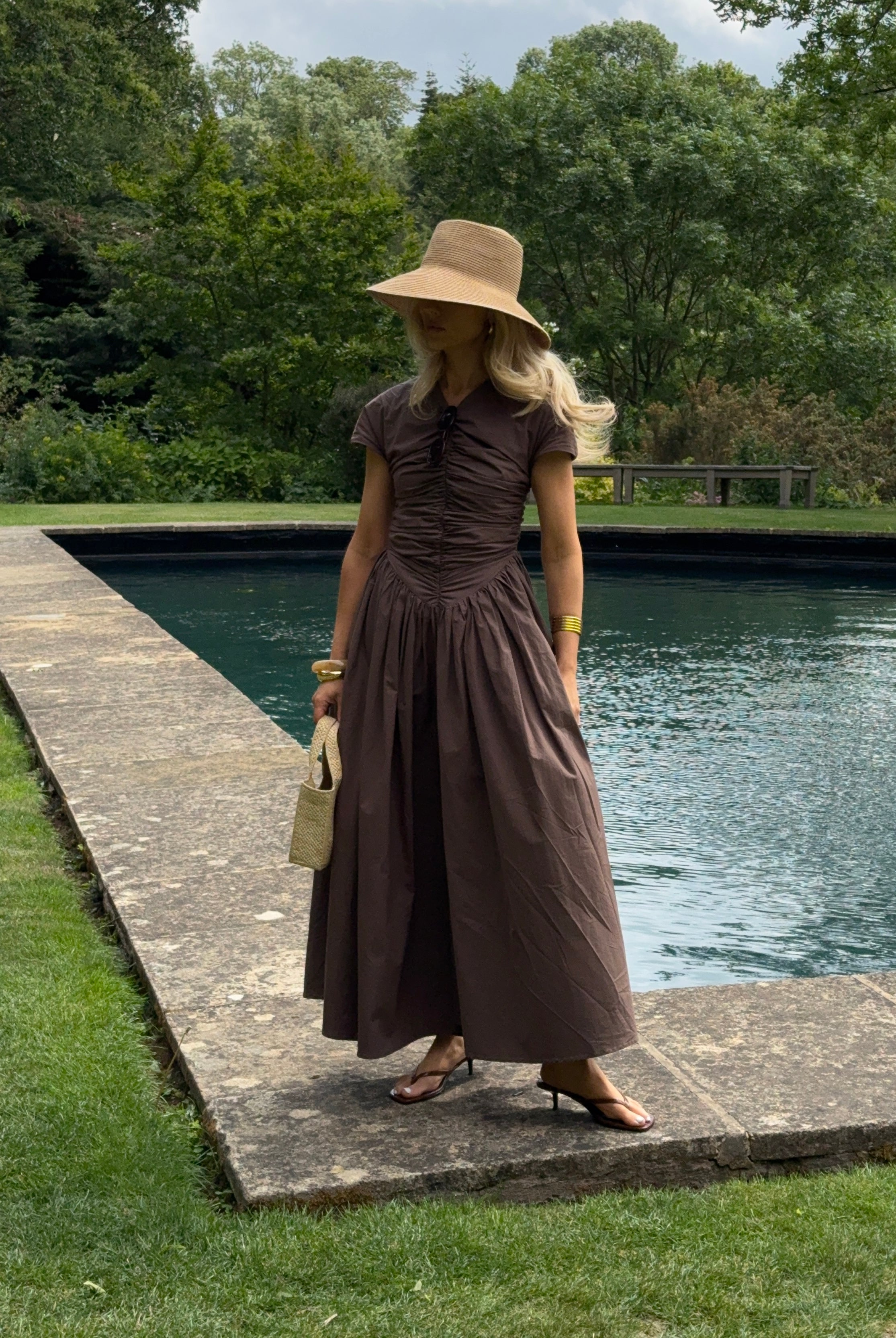 The model wears a brown, short-sleeved ruched dress with a full, flowing ankle-length skirt. She styles the look with a wide-brimmed straw hat, gold bangles, and a small textured handbag, striking a graceful pose in front of a rectangular reflecting pool bordered by a stone walkway and surrounded by lush greenery