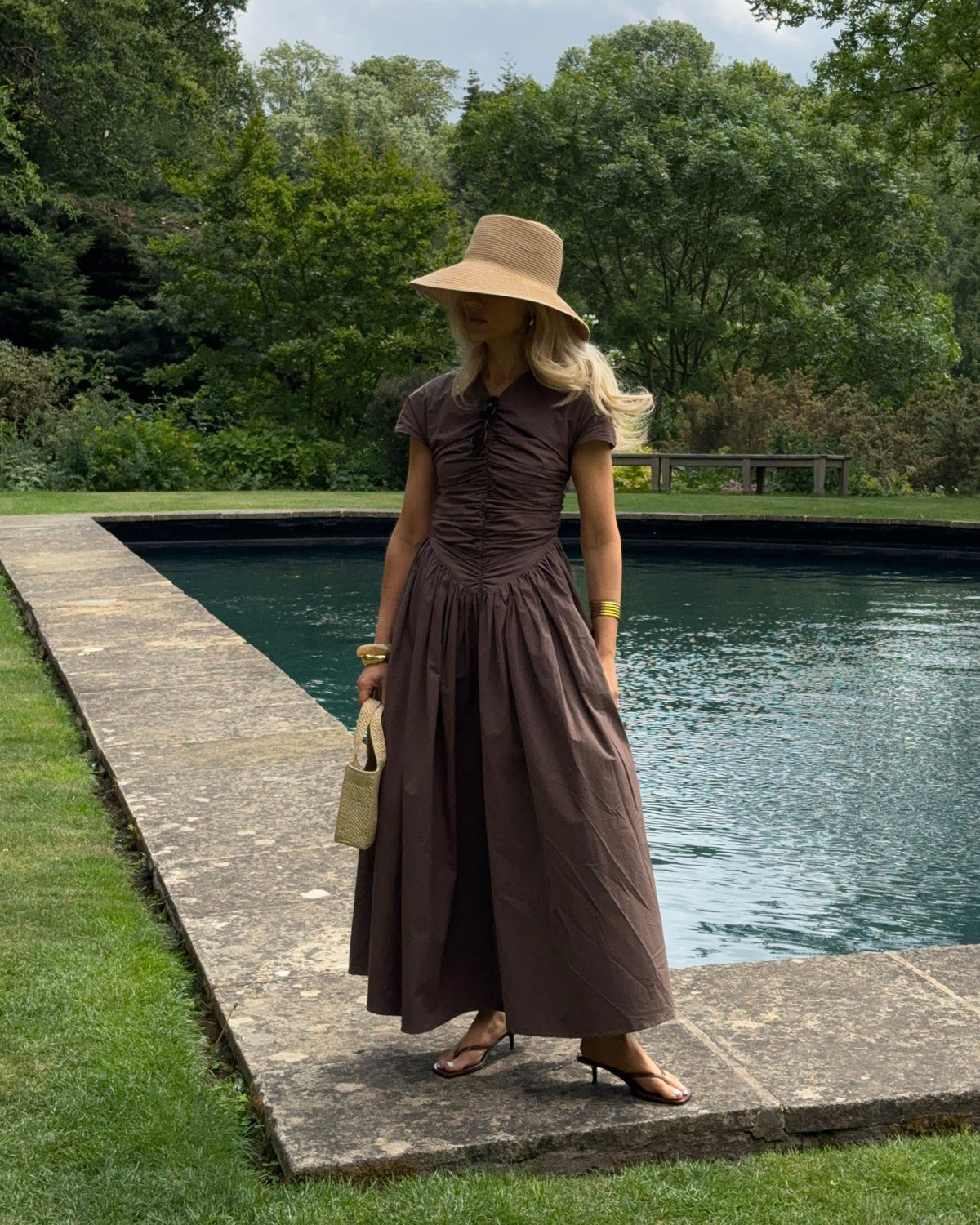 The model wears a brown, short-sleeved ruched dress with a full, flowing ankle-length skirt. She styles the look with a wide-brimmed straw hat, gold bangles, and a small textured handbag, striking a graceful pose in front of a rectangular reflecting pool bordered by a stone walkway and surrounded by lush greenery