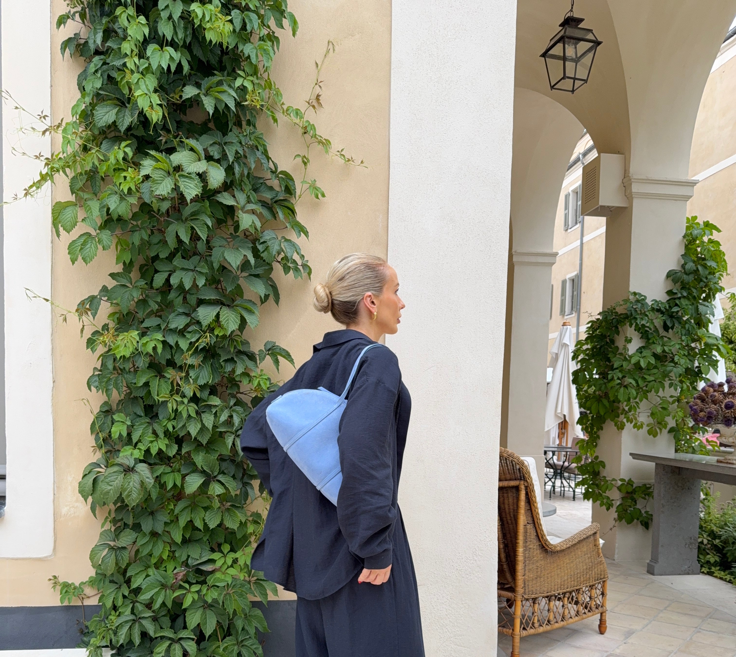 Person wearing a dark blue outfit with a lighter blue handbag in an outdoor setting with a building, plants and chairs in the background