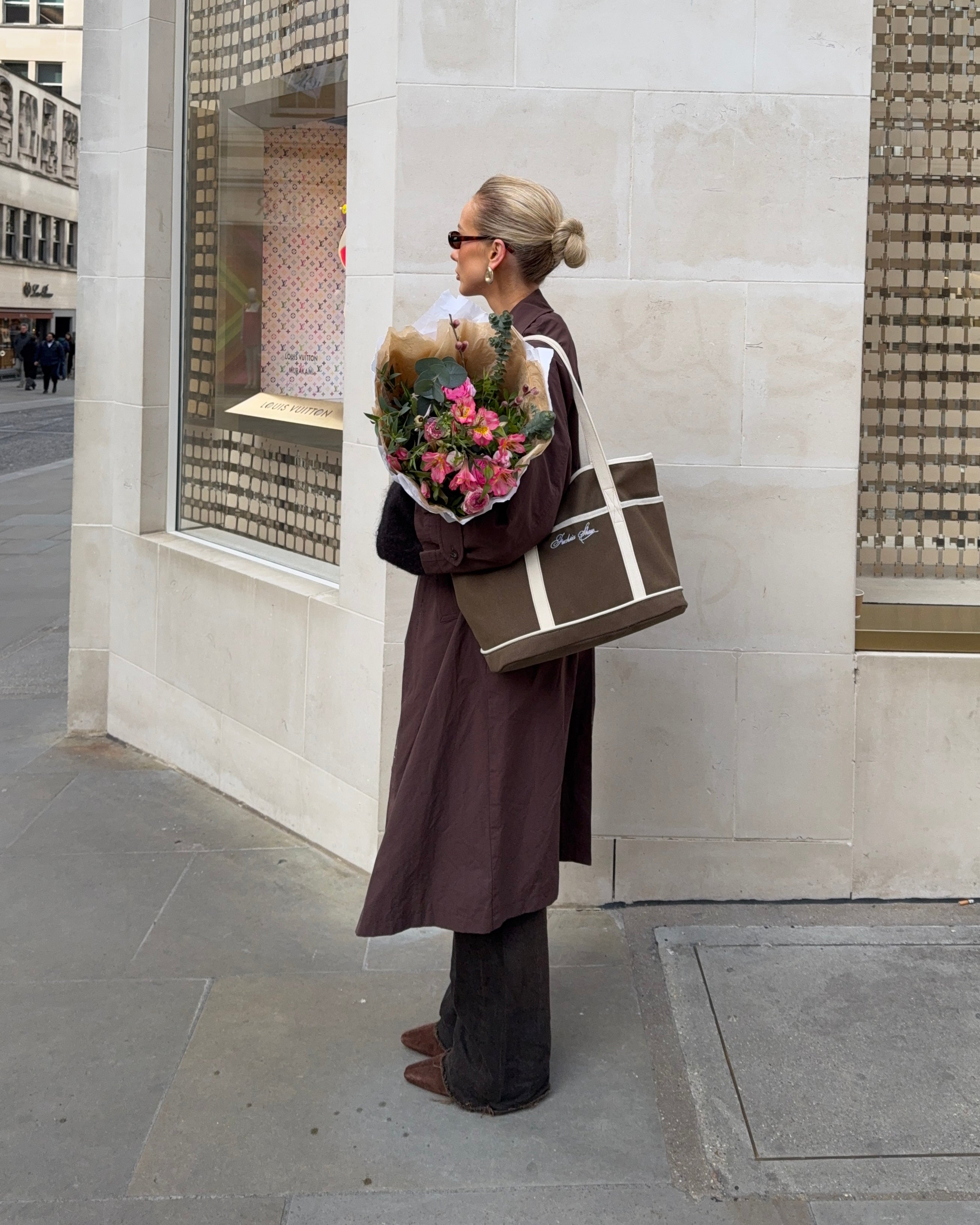 Woman holding some flowers on a street outside a shop wearing a brown outfit with a brown and white bag