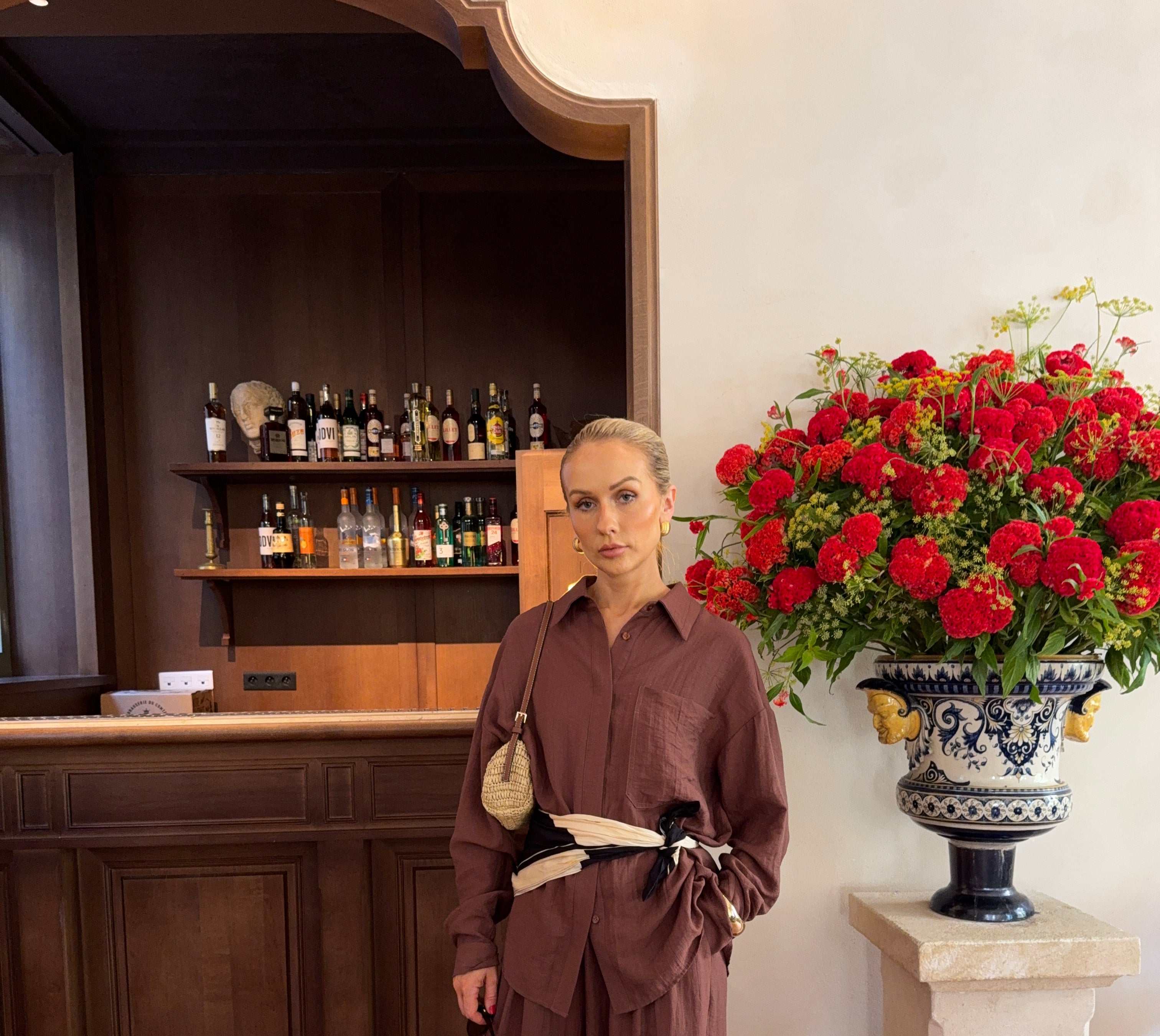 Model in a brown linen shirt and matching trousers accessorised with a scarf bel and handbag, standing in front of a large red and yellow floral arrangement and wooden bar