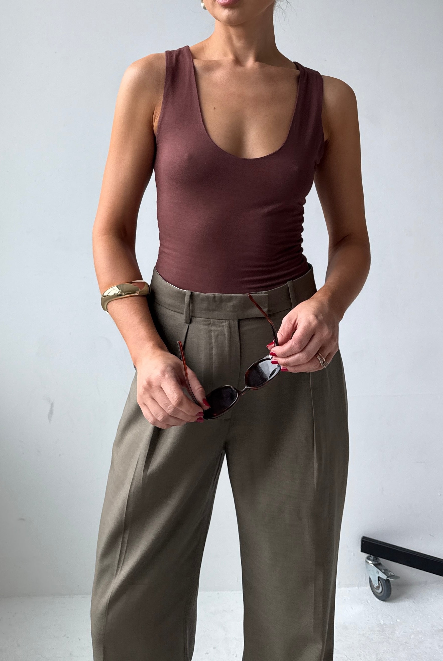 Model wearing a fitting brown tank top and trousers holding sunglasses against a white background