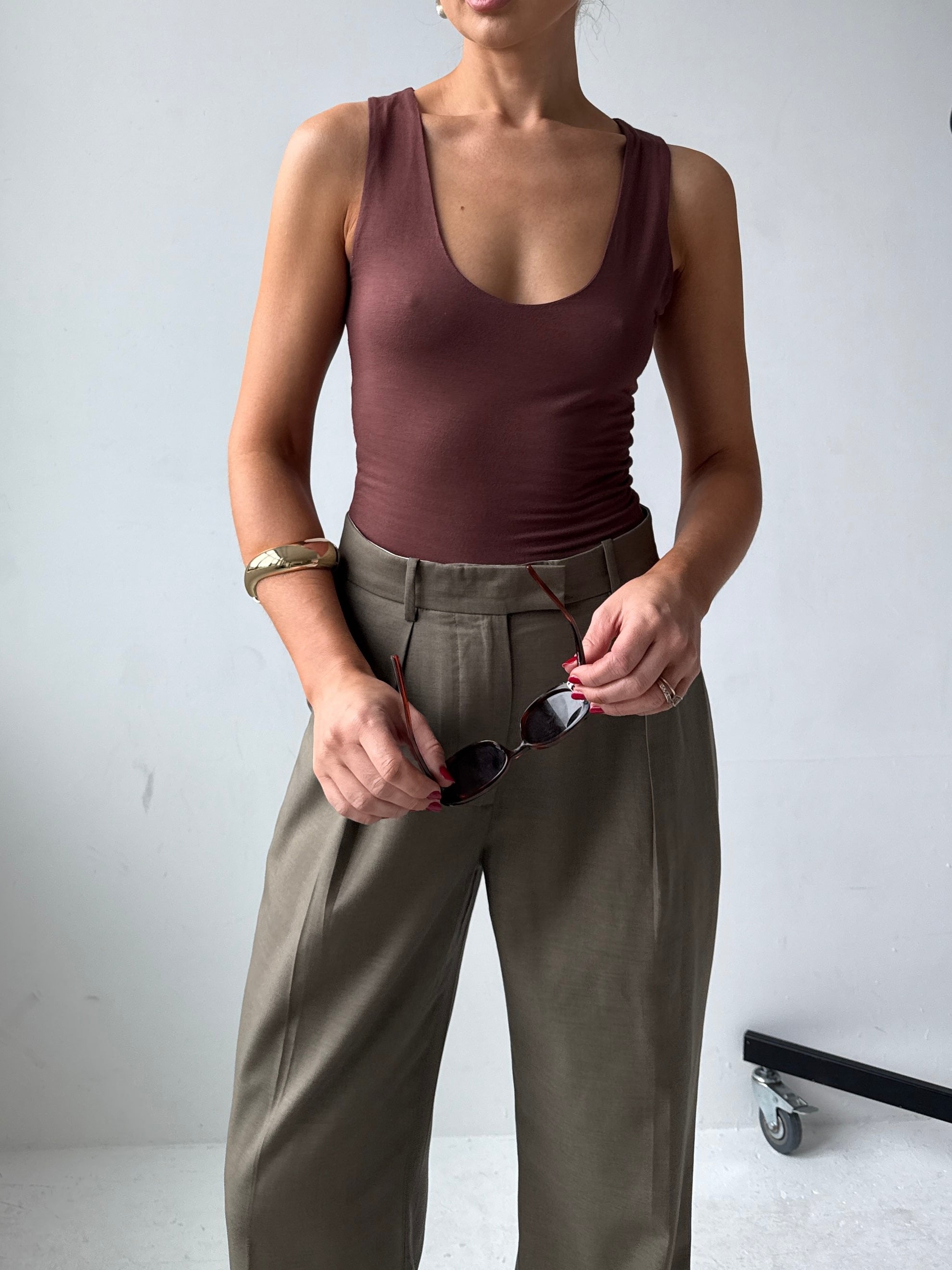 Model wearing a fitting brown tank top and trousers holding sunglasses against a white background