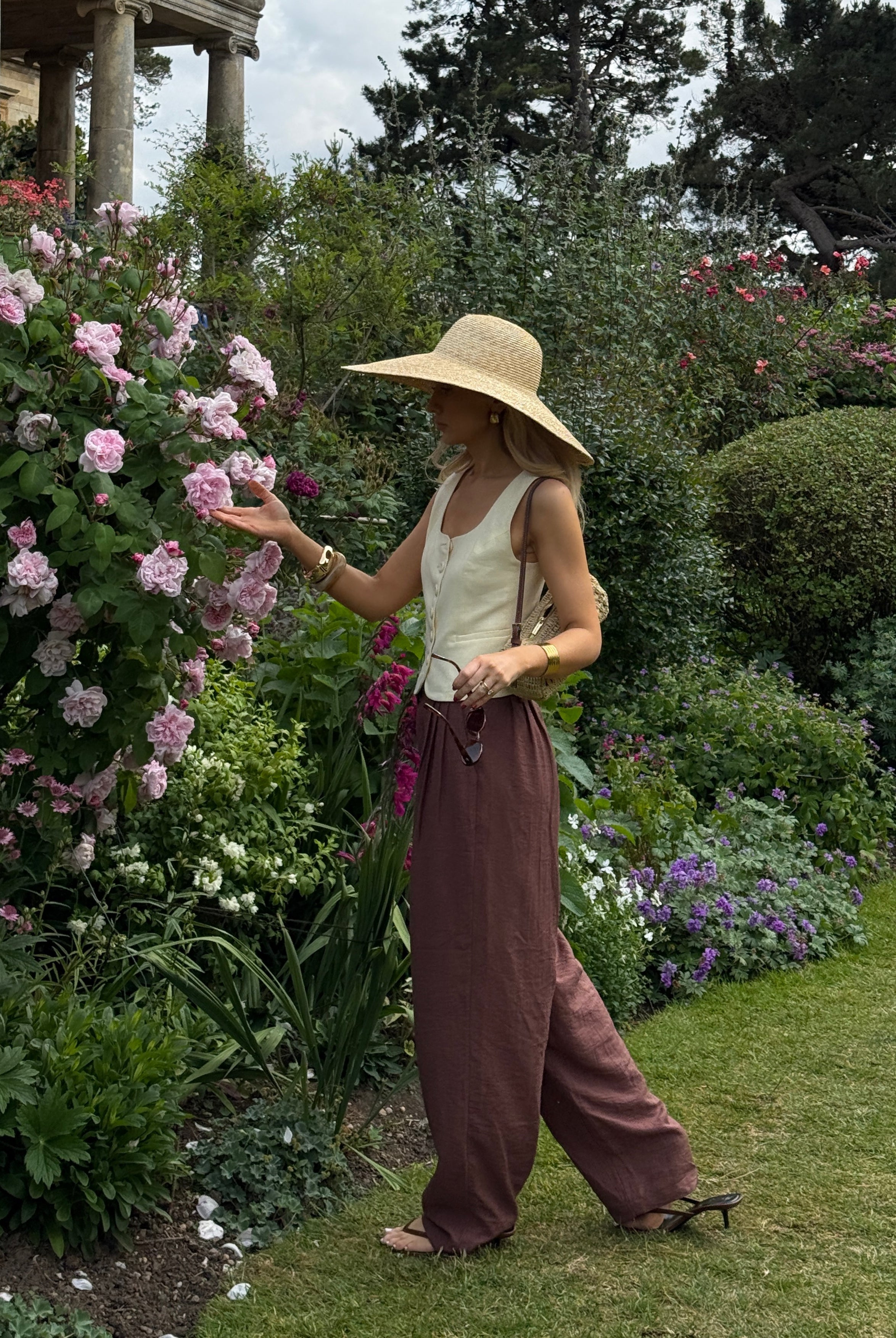 Woman in a vibrant flower garden wearing a wide-brimmed hat and flowing maroon trousers