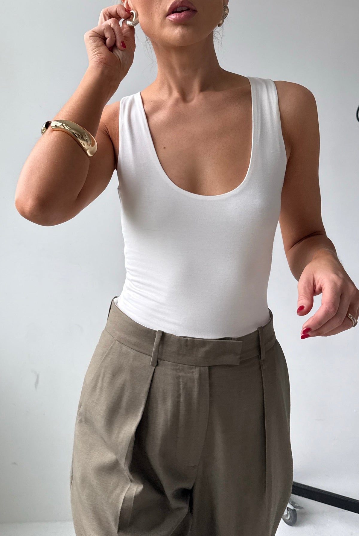 Model wears a fitted white scoop neck tank top with tailored trousers and gold jewellery