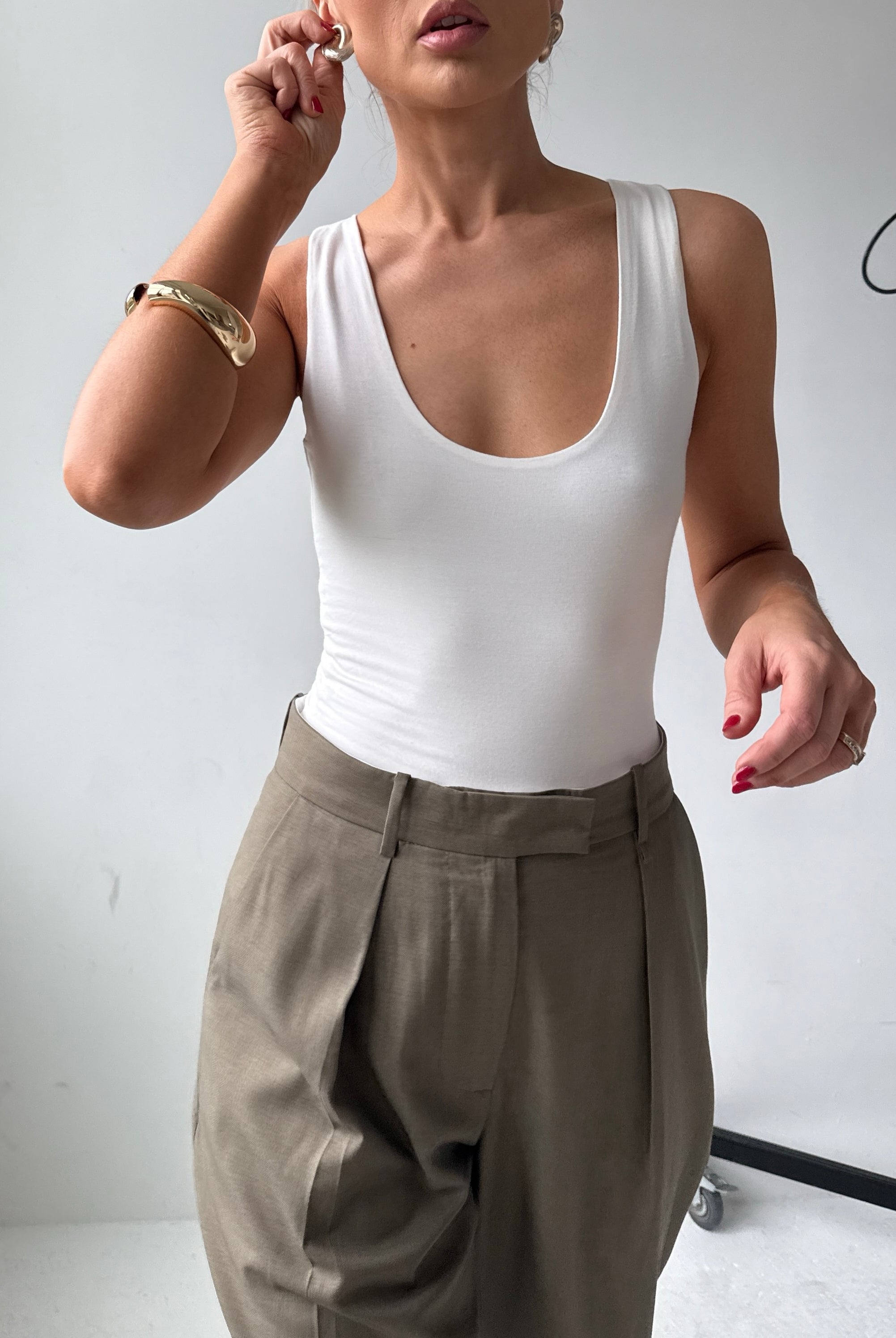 Model wears a fitted white scoop neck tank top with tailored trousers and gold jewellery