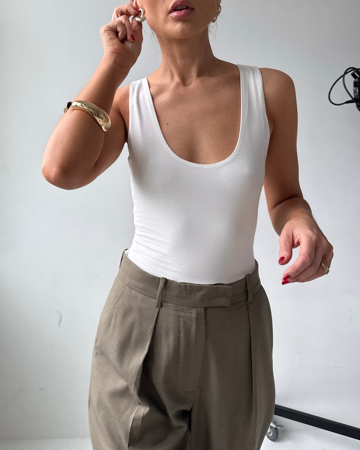 Model wears a fitted white scoop neck tank top with tailored trousers and gold jewellery