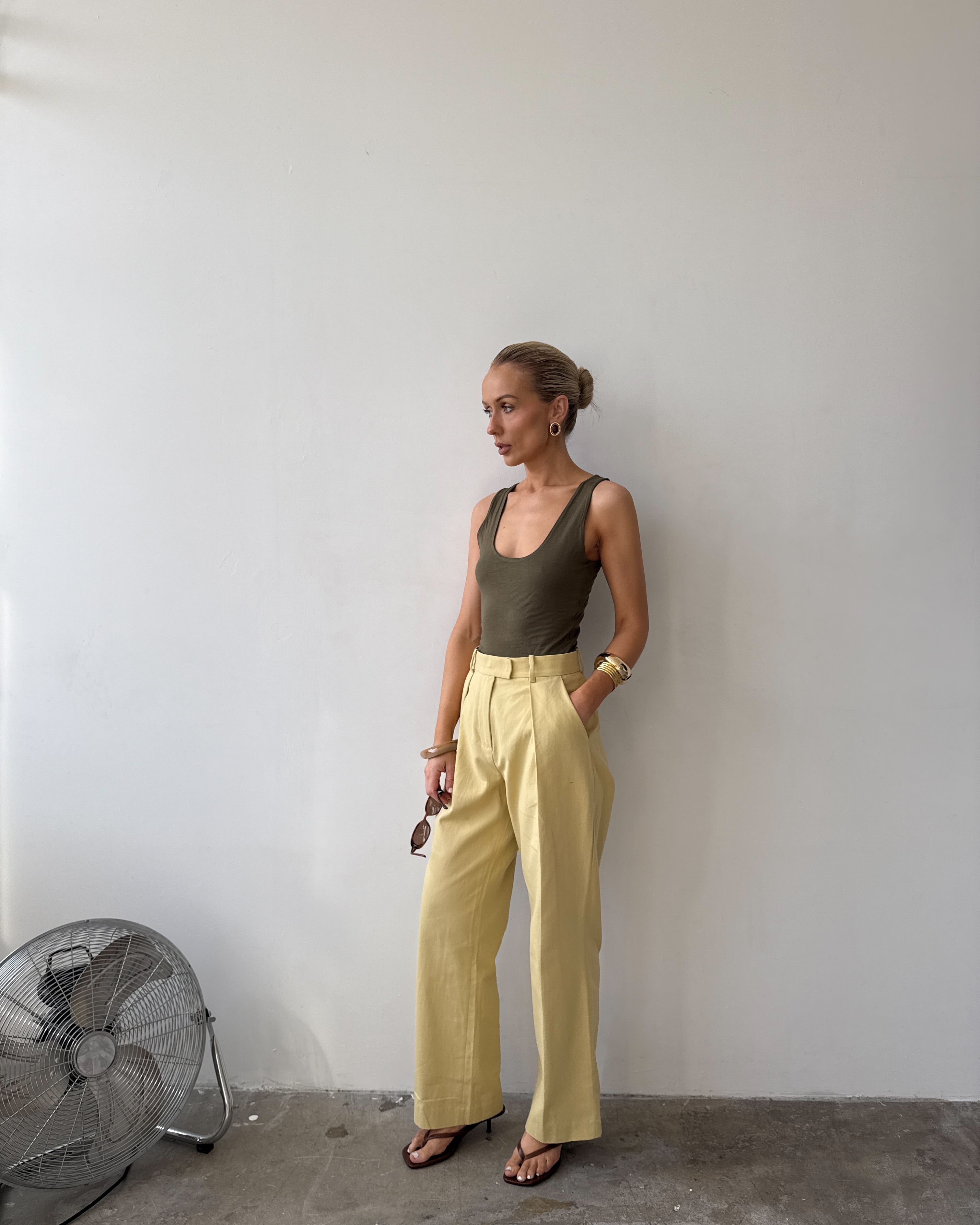Model against a white wall, wearing soft pastel yellow tailored trousers with deep pockets and front pleat styled with a forest green top, with sandals