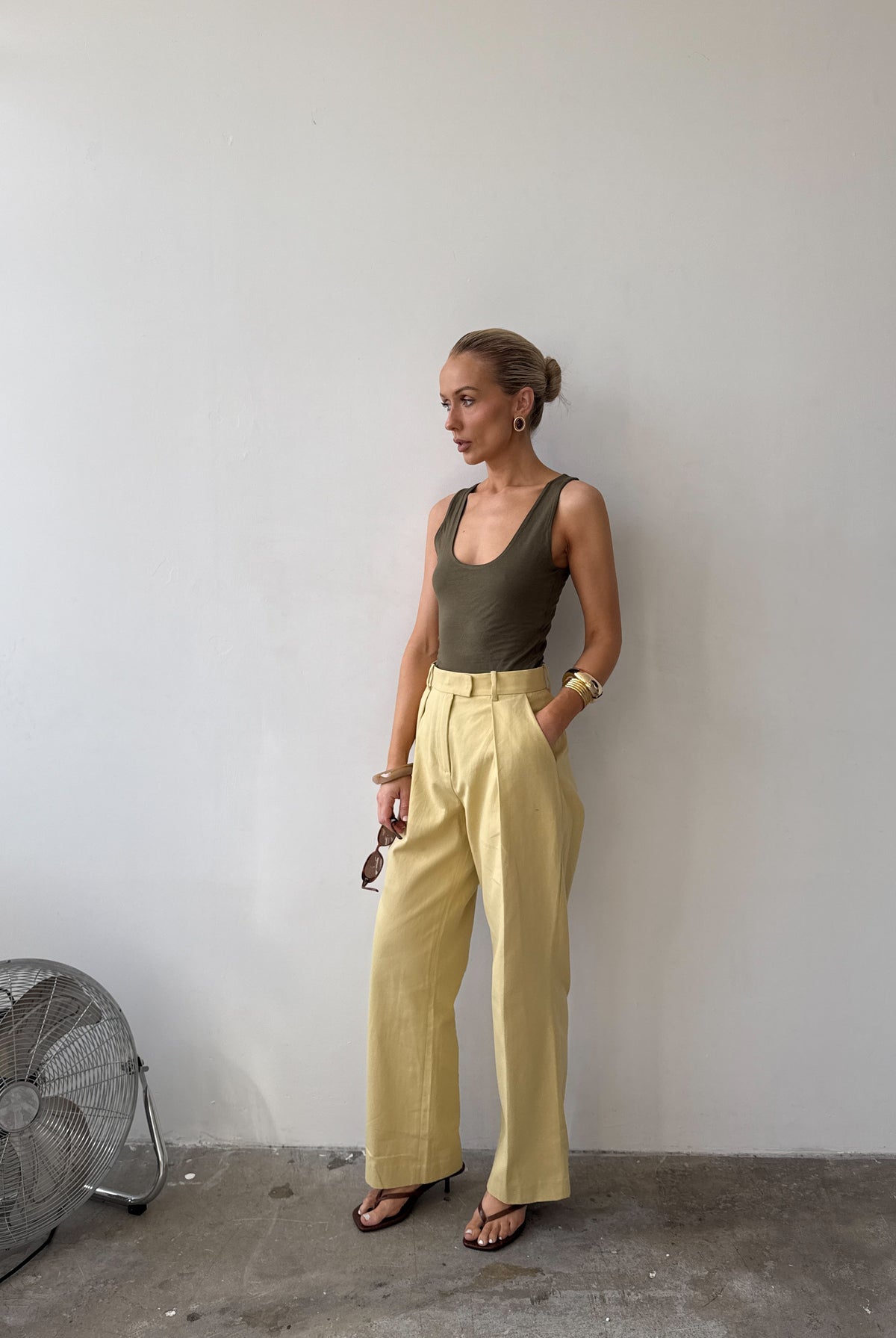 Model against a white wall, wearing soft pastel yellow tailored trousers with deep pockets and front pleat styled with a forest green top, with sandals