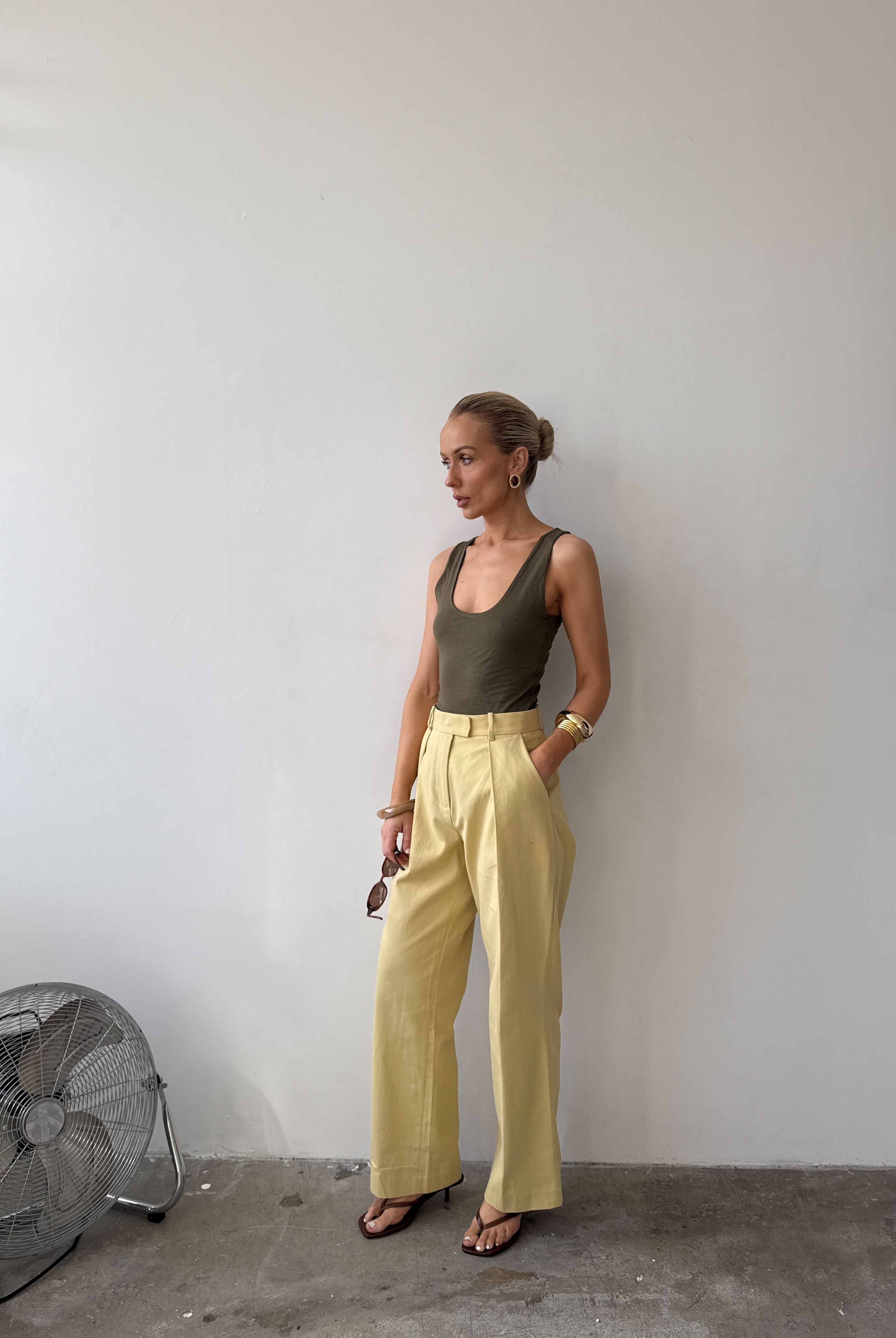 Model against a white wall, wearing soft pastel yellow tailored trousers with deep pockets and front pleat styled with a forest green top, with sandals