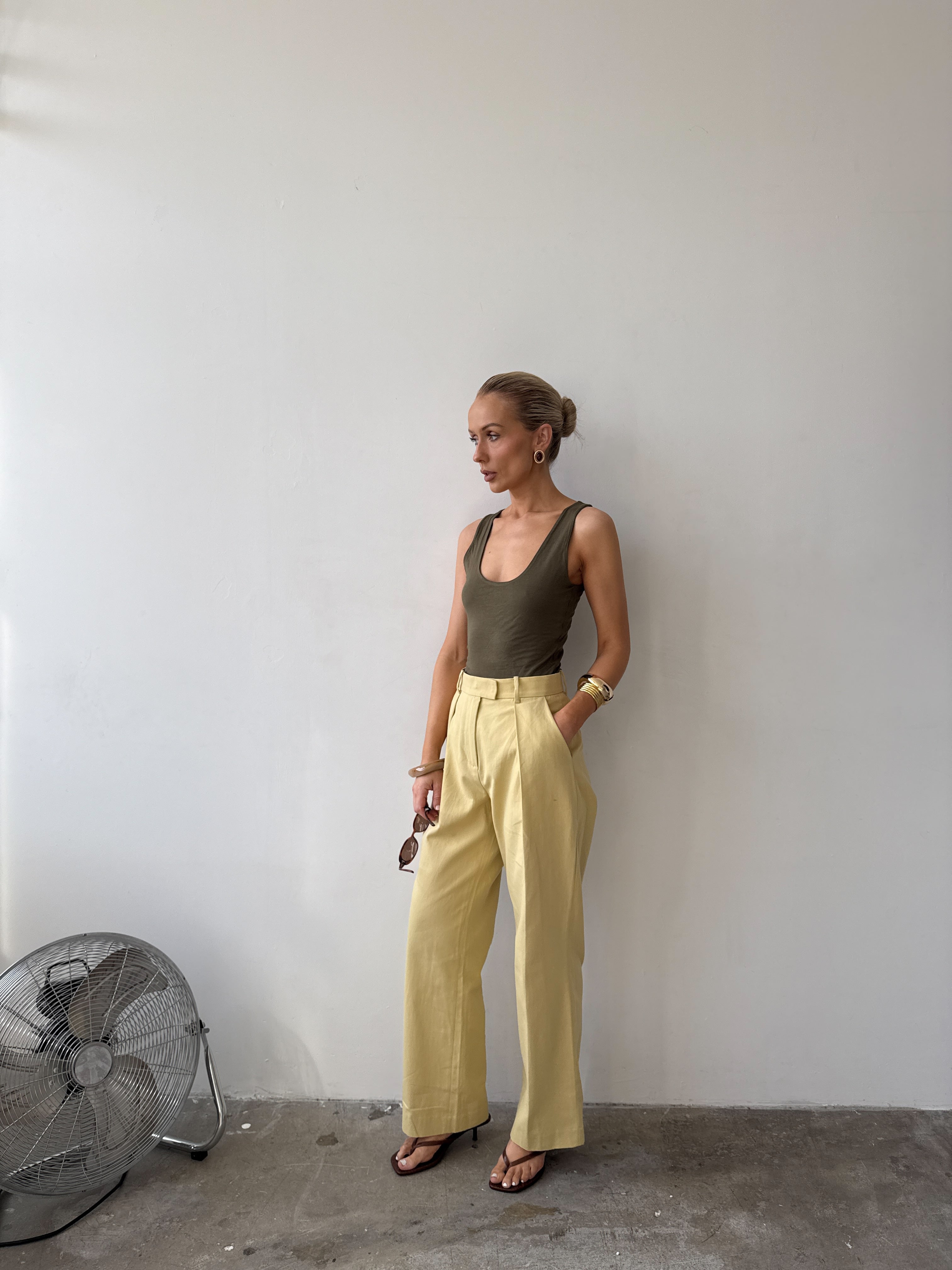 Model against a white wall, wearing soft pastel yellow tailored trousers with deep pockets and front pleat styled with a forest green top, with sandals