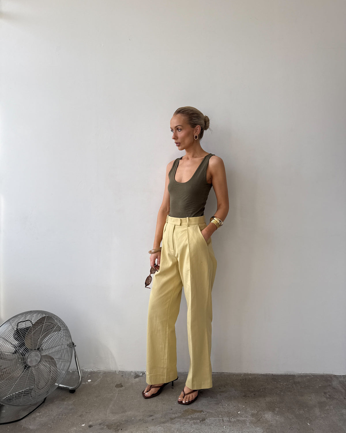 Model against a white wall, wearing soft pastel yellow tailored trousers with deep pockets and front pleat styled with a forest green top, with sandals