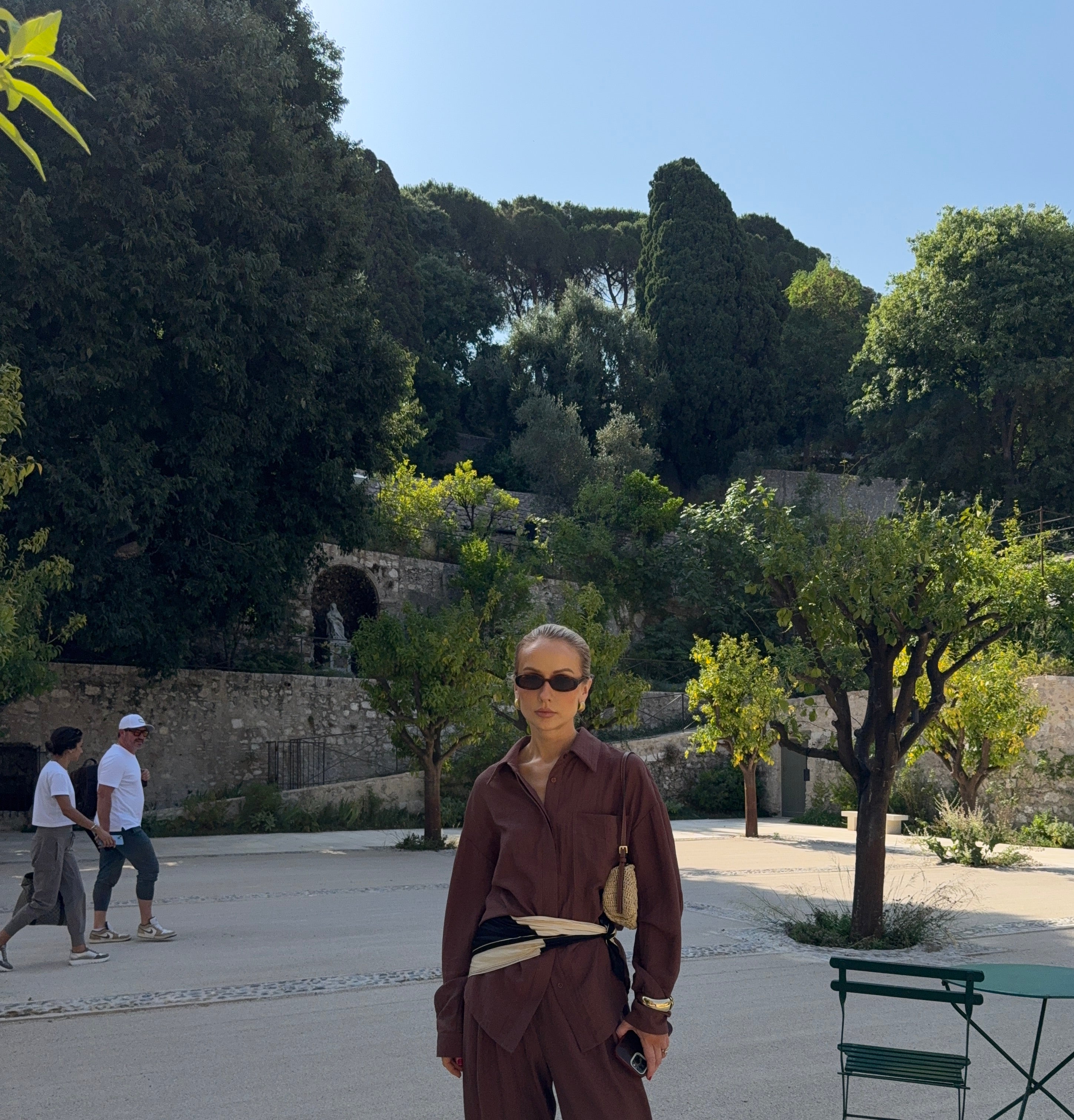 Woman in a brown outfit standing on a paved area with Mediterranean trees and walls in the background