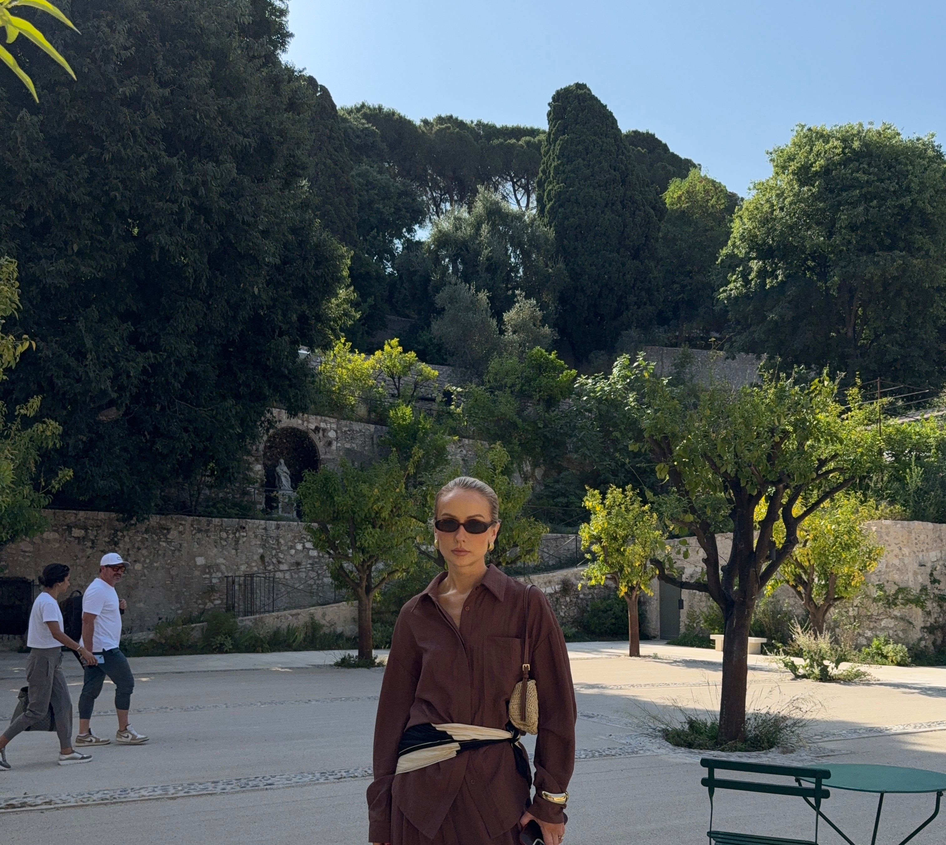 Woman in a brown outfit standing on a paved area with Mediterranean trees and walls in the background