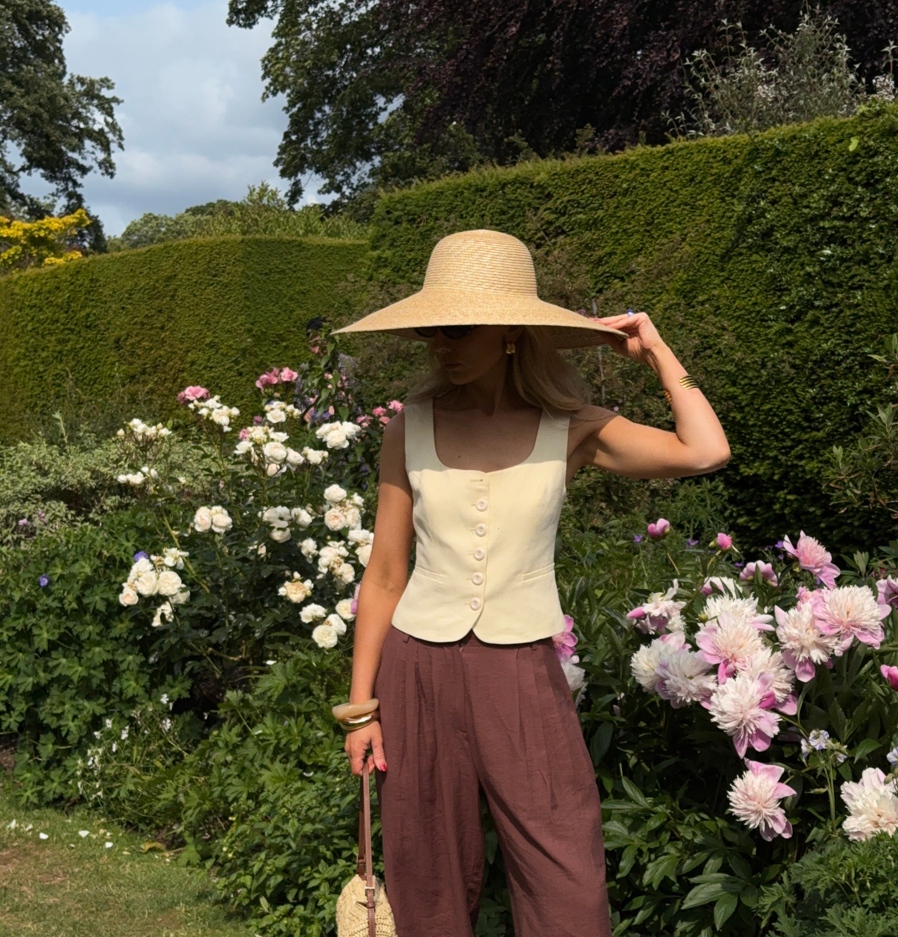 Woman in a sunny garden with box hedges and pale pink flowers flowers, wearing chocolate brown relaxed trousers, a pastel yellow waist coat, and a wide brimmed sunhat with matching handbag