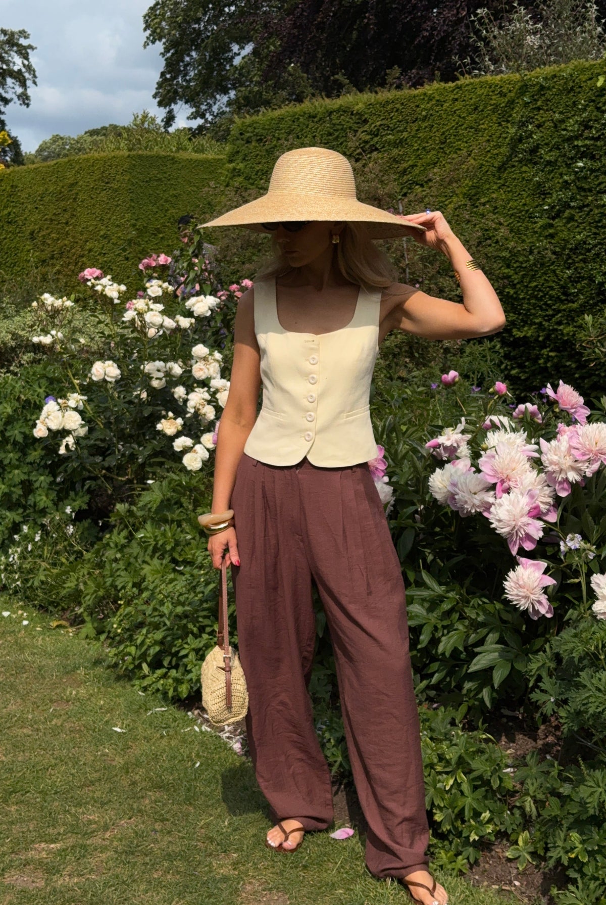 Woman in a sunny garden with box hedges and pale pink flowers flowers, wearing chocolate brown relaxed trousers, a pastel yellow waist coat, and a wide brimmed sunhat with matching handbag