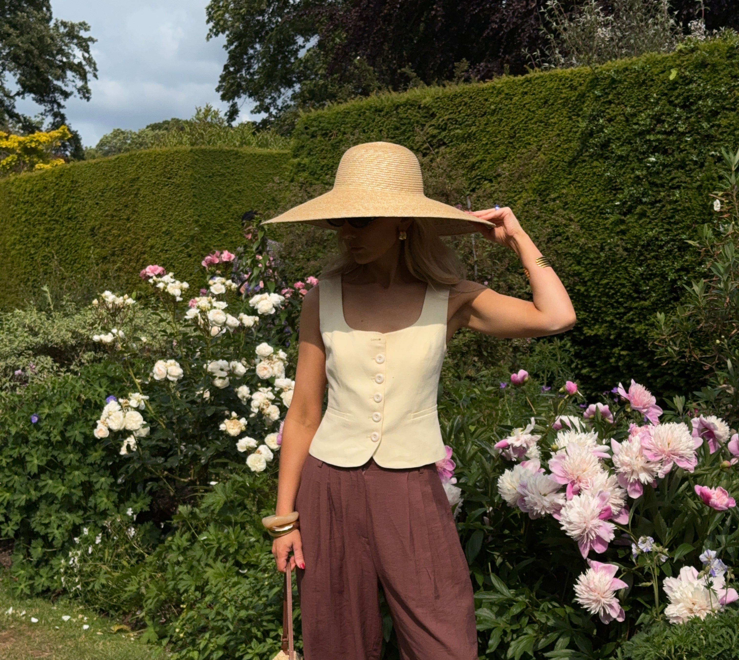 Woman in a sunny garden with box hedges and pale pink flowers flowers, wearing chocolate brown relaxed trousers, a pastel yellow waist coat, and a wide brimmed sunhat with matching handbag