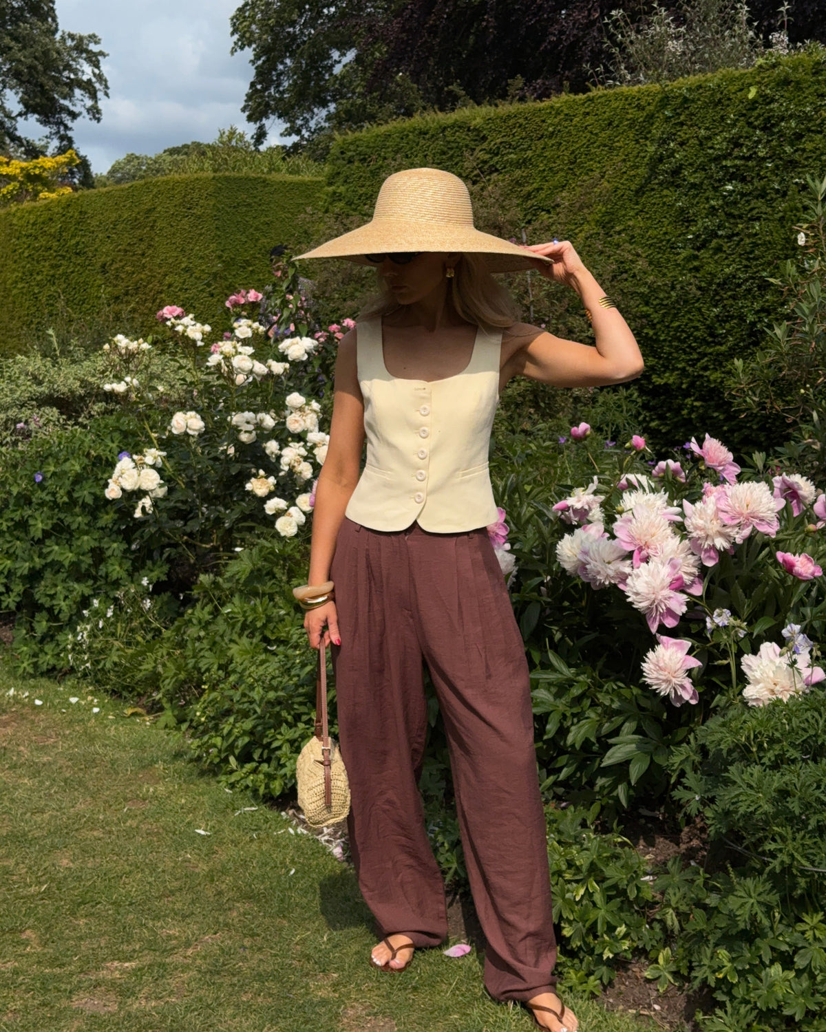 Woman in a sunny garden with box hedges and pale pink flowers flowers, wearing chocolate brown relaxed trousers, a pastel yellow waist coat, and a wide brimmed sunhat with matching handbag
