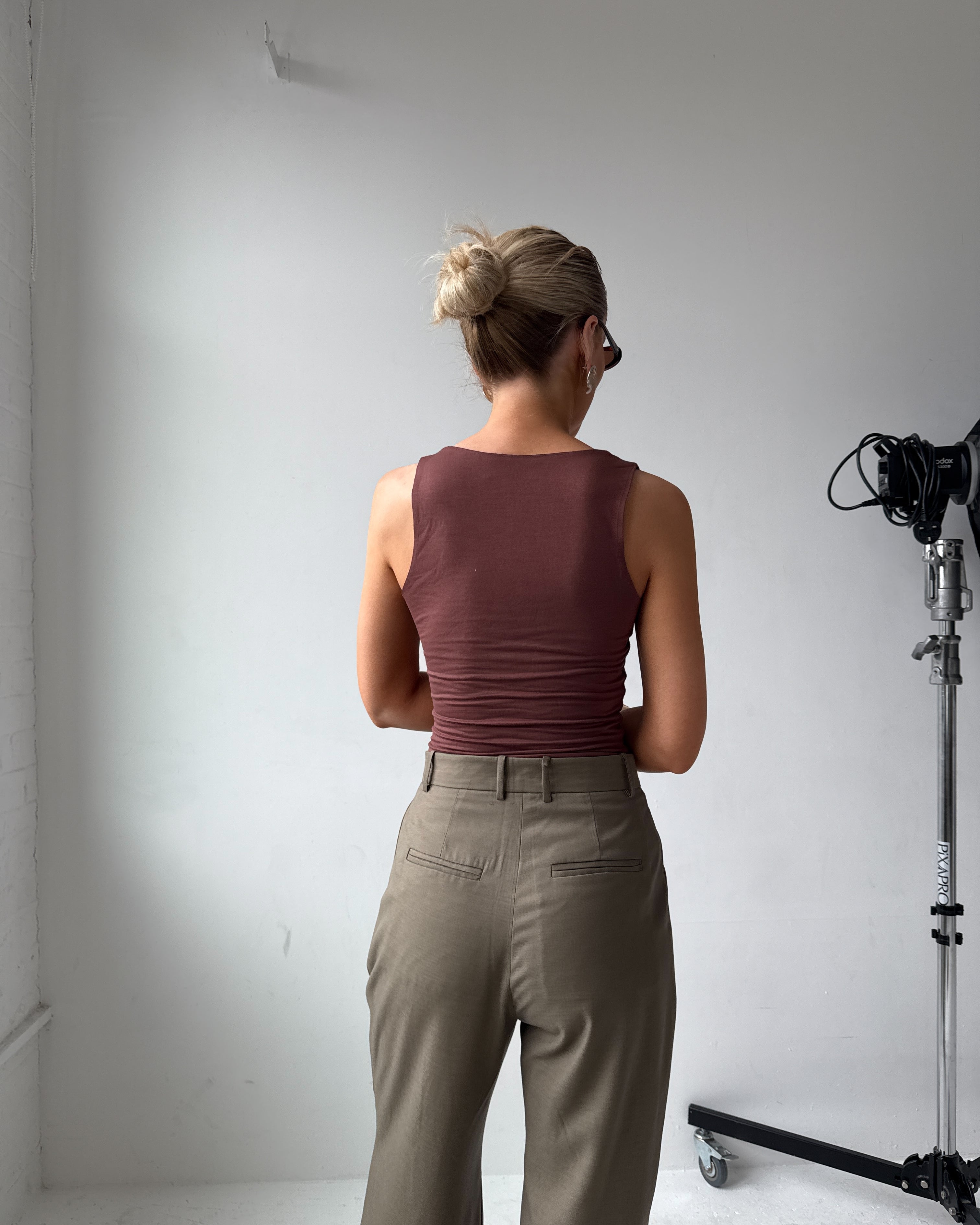 Model wearing a maroon top and beige-grey trousers standing in a studio setting with camera equipment
