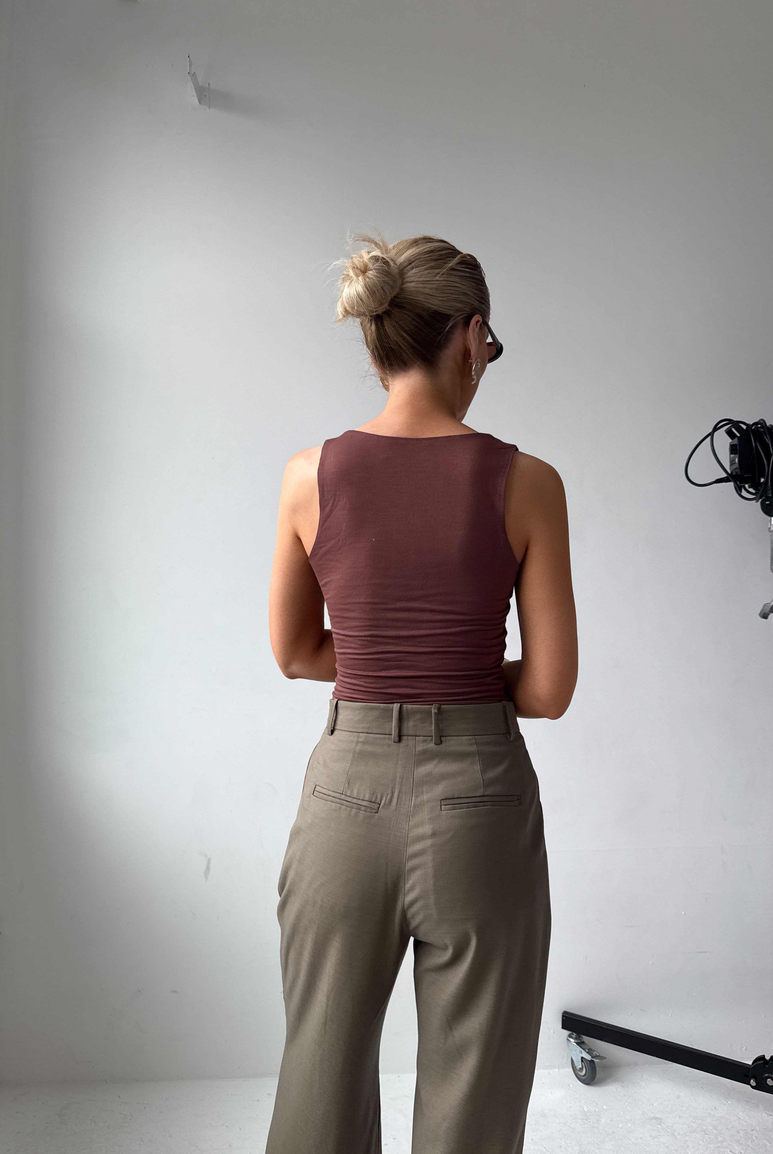 Model wearing a maroon top and beige-grey trousers standing in a studio setting with camera equipment