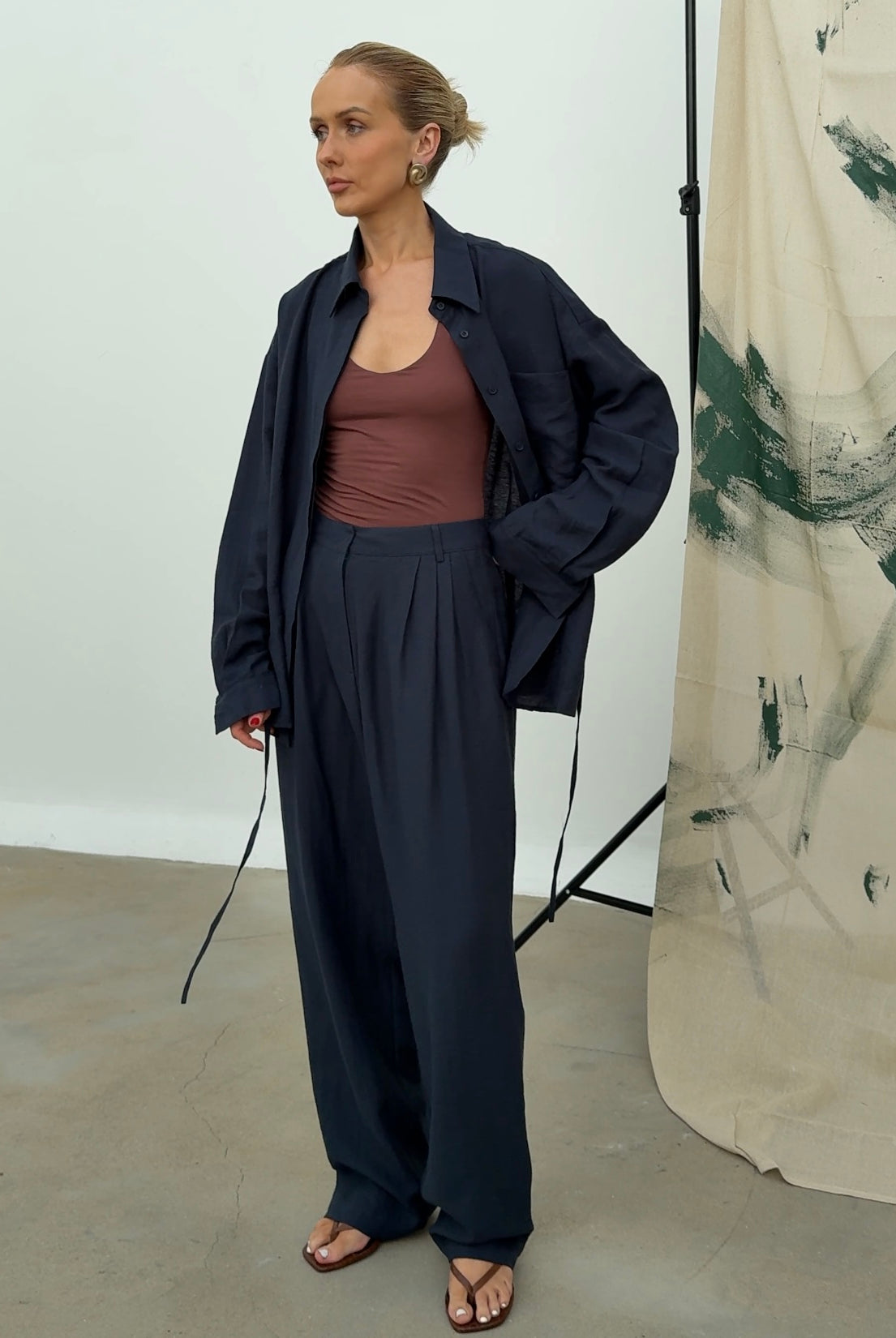Model wears a navy linen shirt paired with matching trousers, standing in a minimalist room with an abstract painted background